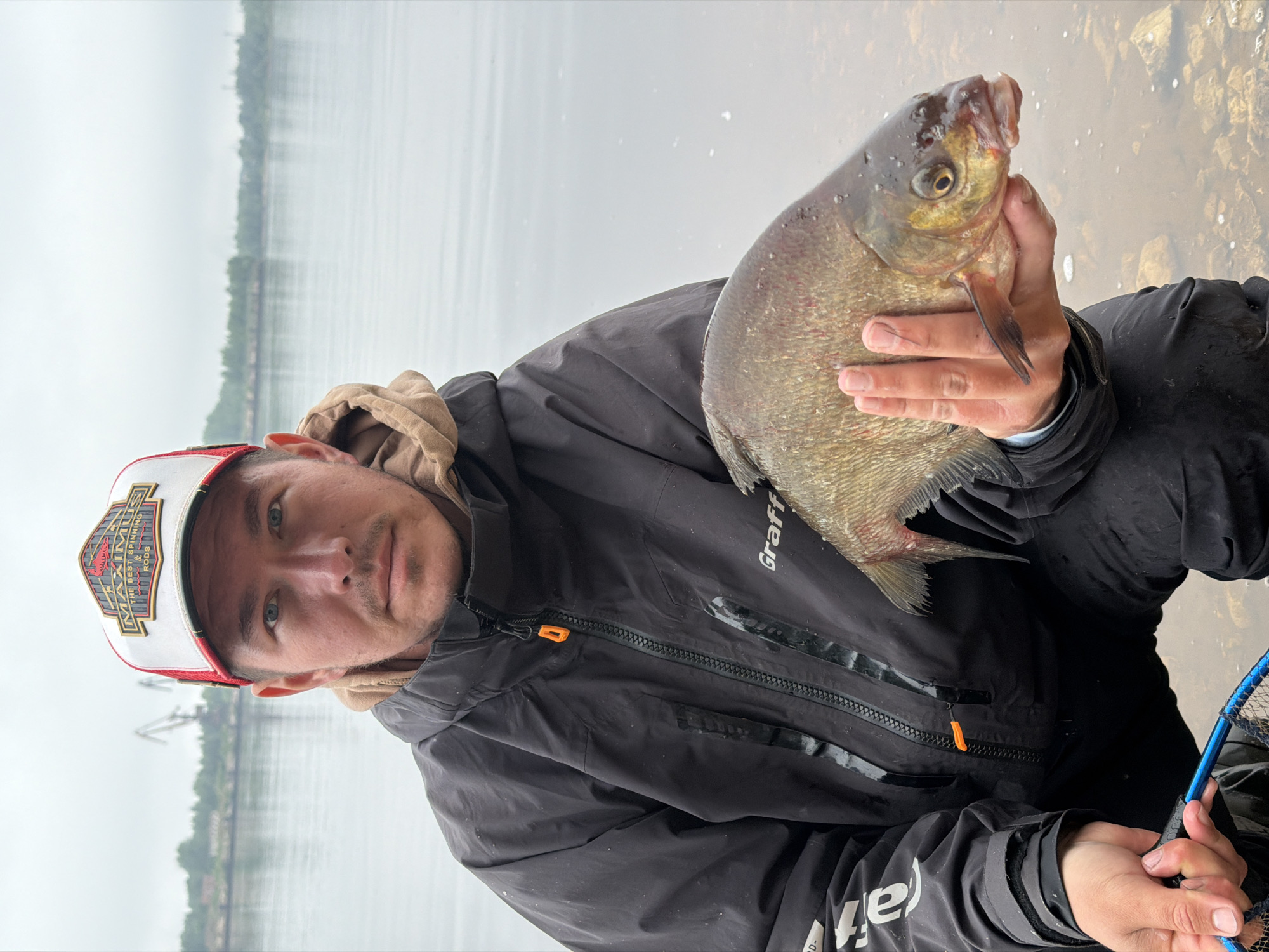 A male angler is depicted holding a large, freshly caught fish by a wide body of water in Chaikovsky, Russia. The man, looking directly at the camera with a neutral expression, wears a black waterproof jacket with the "Graff" brand logo visible on his right shoulder area. Underneath, he has a beige hooded sweatshirt. On his head, he wears a white and red baseball cap featuring a prominent emblem with the text "MAXIMUS THE BEST SPINNING & RODS".
He holds a broad-bodied, silvery-brown fish, resembling a bream, in his right hand. The fish displays distinct scales, a large eye, and a small, slightly upturned mouth. Its fins are clearly visible. A portion of a blue fishing net, including its handle and rim, is visible in the bottom right corner of the frame, held in his left hand.
The background features a vast expanse of calm, grey water, suggesting a large lake or river like the Kama River in Chaikovsky. The sky is overcast, contributing to the soft, diffused lighting and the subdued reflections on the water's surface. A distant green shoreline, densely covered with trees, runs horizontally across the mid-ground. Faint outlines of what appear to be industrial structures, possibly cranes, can be seen on the far shore. The scene conveys the quiet satisfaction of a successful fishing outing on a cloudy day.