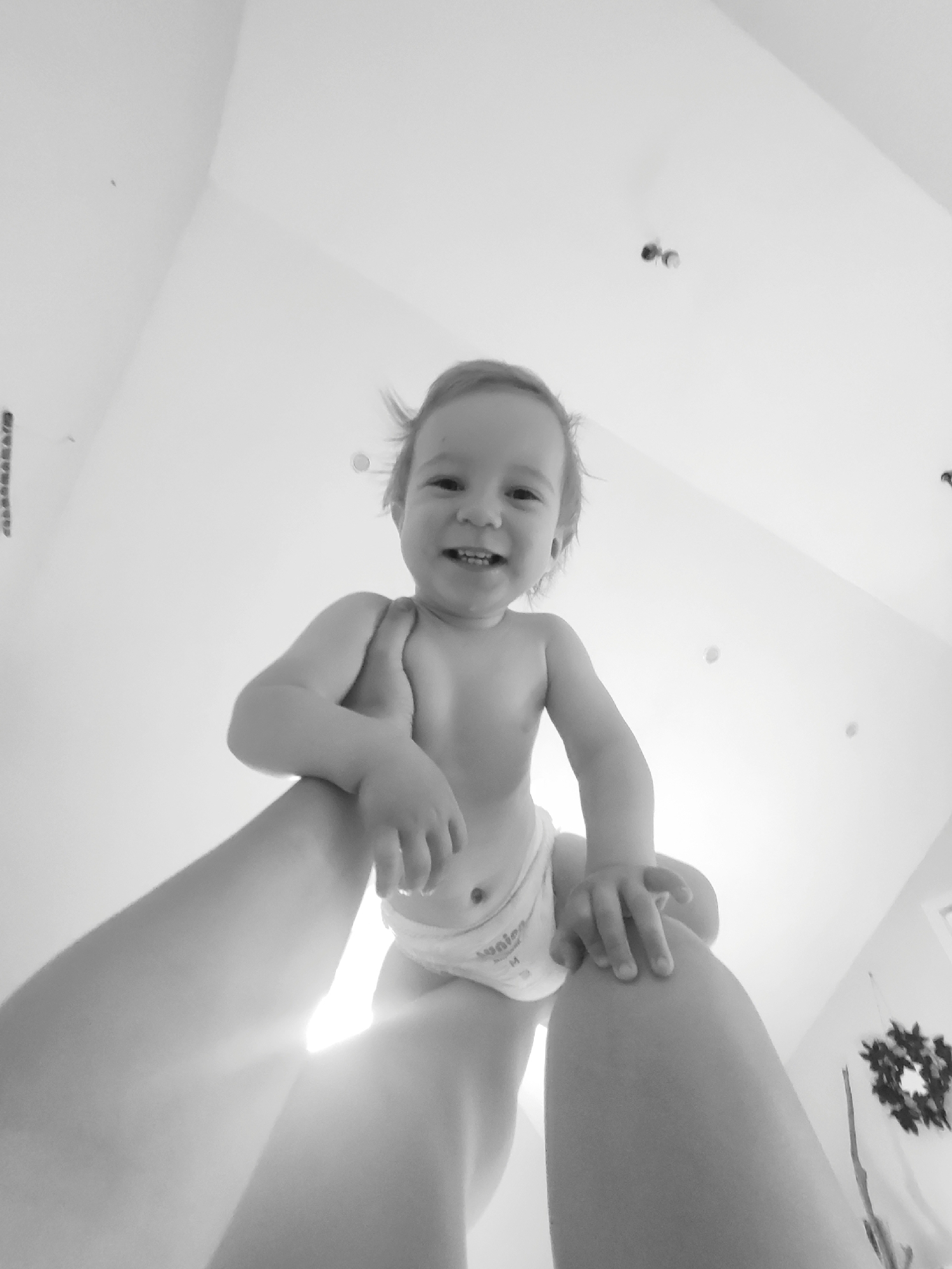 A grayscale, low-angle shot captures a baby being held up, presumably by a parent or caregiver. The baby is wearing a diaper and has a cheerful expression. The scene appears to be indoors, with a white ceiling and walls, as well as a wreath visible. The location is not identifiable.