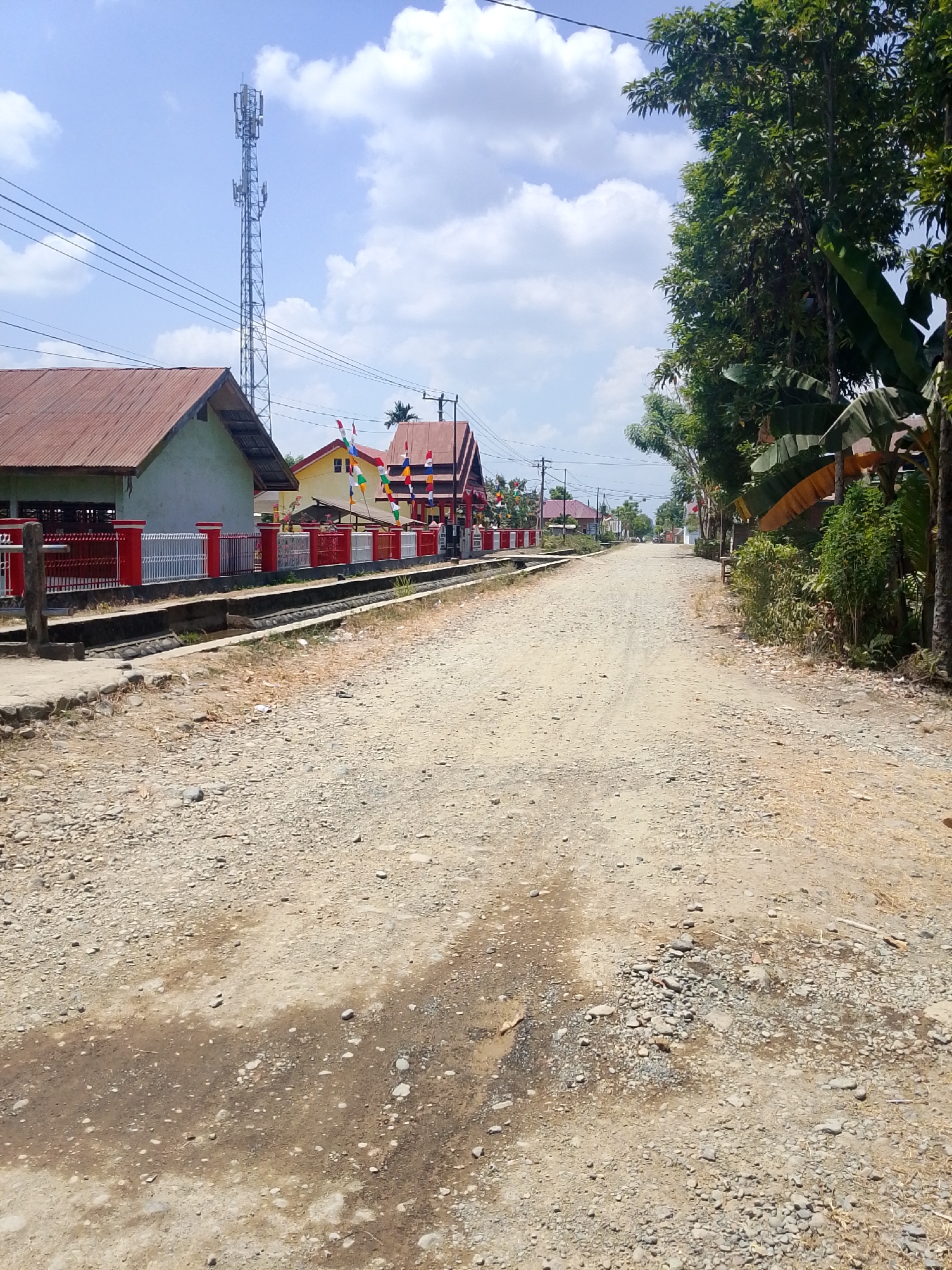 The image depicts a dusty, gravel road in Se'pon, Indonesia, with buildings and vegetation lining its sides. A tall cell tower stands prominently against a bright blue sky dotted with white clouds on the left side of the frame. Adjacent to the road are several buildings, including one with a reddish-brown corrugated metal roof and a white and red fence. Further down the road, other structures with colorful flags are visible. On the right side of the road, dense green trees and foliage create a natural barrier, with some banana leaves also present. The road itself is rough and uneven, with scattered rocks and patches of damp earth. No people or vehicles are visible in the scene.