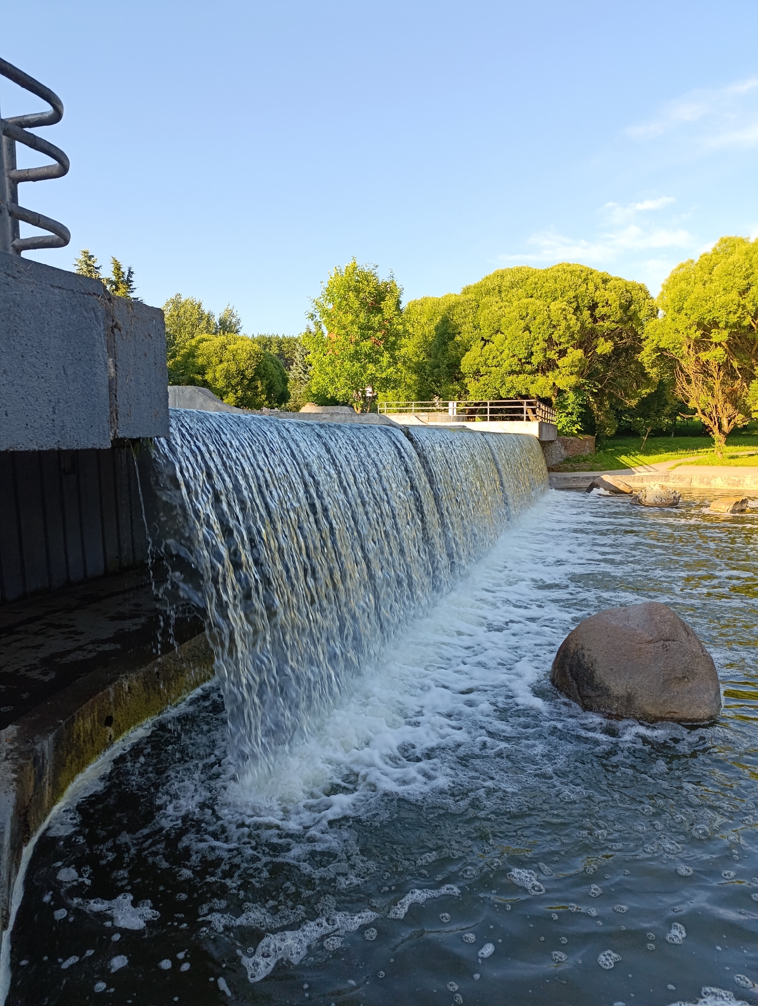 The image shows a waterfall in a park with a clear sky. A large concrete structure with a metal railing is visible on the left, with water flowing over the edge to create a curtain of falling water. The water below is agitated, with white foam and bubbles. A large, rounded rock is partially submerged in the water on the right side of the frame. In the background, lush green trees line the park, with a wooden bridge and railing visible further back. The overall scene suggests a tranquil and natural setting within a city. This waterfall is located in Gorky Park in Minsk, Belarus.