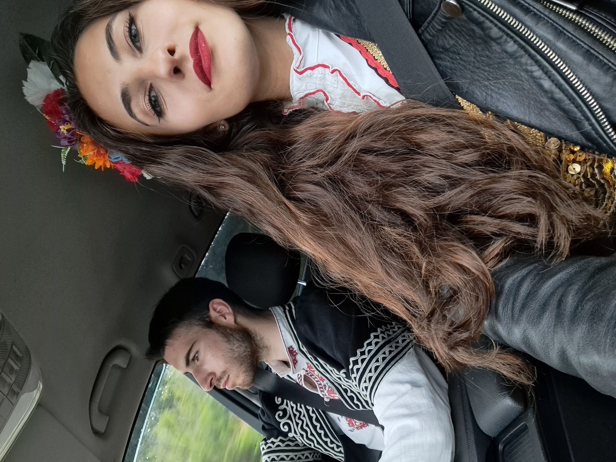 Two individuals are in a car. The female, positioned in the upper portion of the frame, has long brown hair, wears a flower crown, and a red and white ruffled top. She is leaning against a black leather jacket. The male, located in the lower portion of the frame, is wearing a white shirt with embroidered black and white patterns. They are inside a car, with the interior roof and side window visible. The external environment appears to be a blur of green vegetation, suggesting they are driving. The city is unknown.