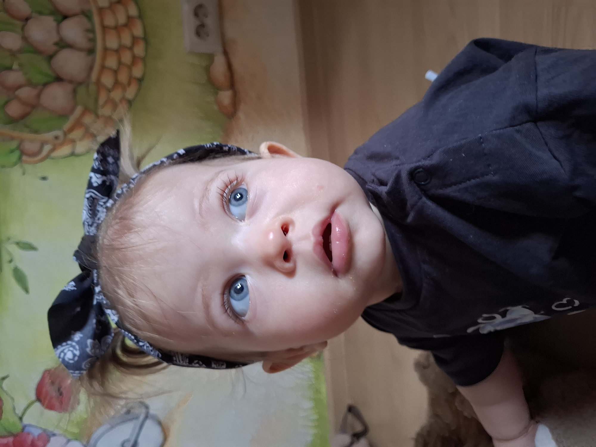 An indoor, eye-level shot, rotated 90 degrees clockwise, features a baby in Chaikovsky, Russia. The baby, with light-colored hair, fair skin, and piercing blue eyes, is the central subject. They wear a dark blue or black long-sleeved top and a patterned bandana (black with white paisley-like designs) tied on their head. The baby's mouth is slightly open, and there is visible moisture around the lips and nose, suggesting drool. The expression is one of curiosity, with the baby looking upwards and slightly to the right.
The background consists of a wall adorned with a colorful mural or wallpaper. To the left (when correctly oriented), a large woven basket filled with brown, rounded shapes (possibly mushrooms or fruit) is depicted, alongside green foliage. Below the basket, red elements, resembling berries or apples, are visible. Further to the right on the wall, a white electrical outlet is mounted. Below the baby, a light brown, possibly wooden or laminate, surface can be seen. No specific time of day or weather conditions are discernible. There is no visible text in the image.