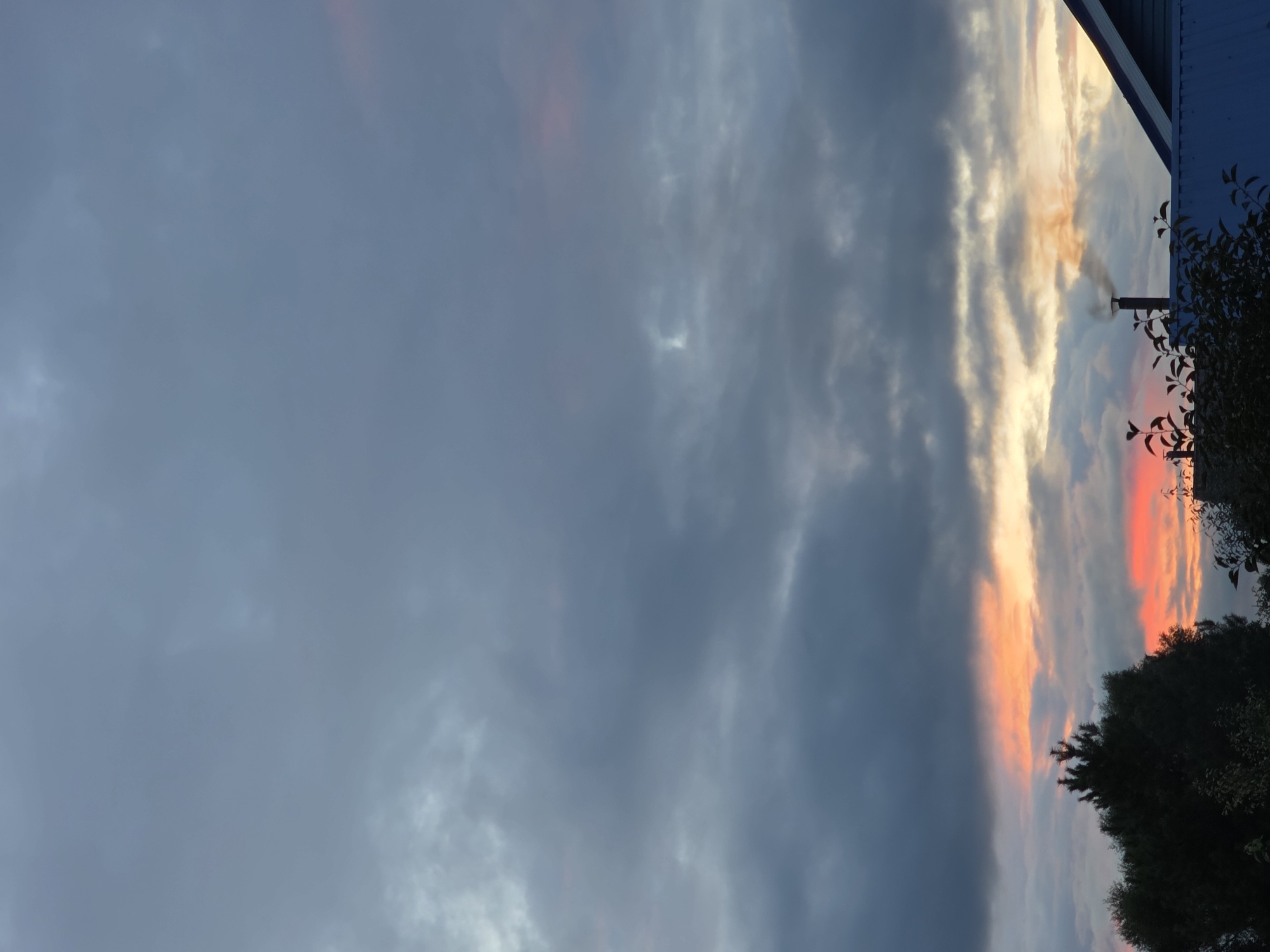The image depicts a cloudy sky with streaks of orange and pink, suggesting a sunset or sunrise, over a residential area. In the foreground, silhouetted trees and the roofline of a blue-sided house are visible. A chimney on the house is emitting a plume of dark smoke, indicating an active fire. The setting appears to be a rural or suburban environment, possibly in the city of Rechytsy, Russia, given the context provided. There are no people or discernible actions depicted in the scene.