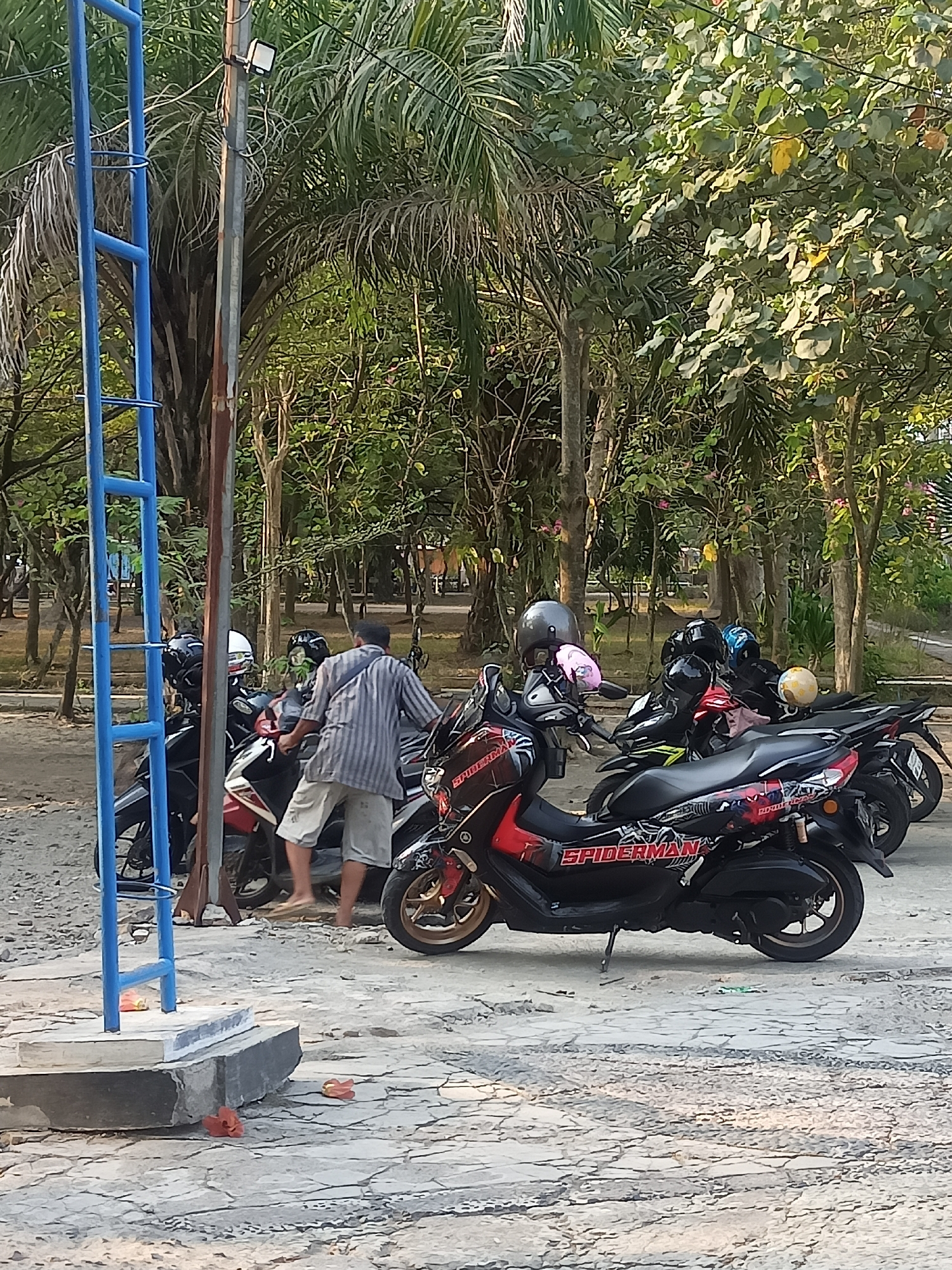 In the city of Girimargo, Indonesia, an outdoor scene captures a group of parked motorcycles and a person. A blue metal ladder-like structure stands on the left, adjacent to a utility pole with a mounted light fixture. A person wearing a striped short-sleeved shirt and light-colored shorts stands between two motorcycles, facing away from the camera. Several motorcycles are parked, with one prominent red and black scooter featuring "SPIDERMAN" graphics. Other motorcycles and helmets are also visible. The ground is paved with cracked concrete, and scattered orange flower petals are present on the ground and on the base of the ladder structure. Lush green trees with dense foliage dominate the background, suggesting a park or roadside setting.