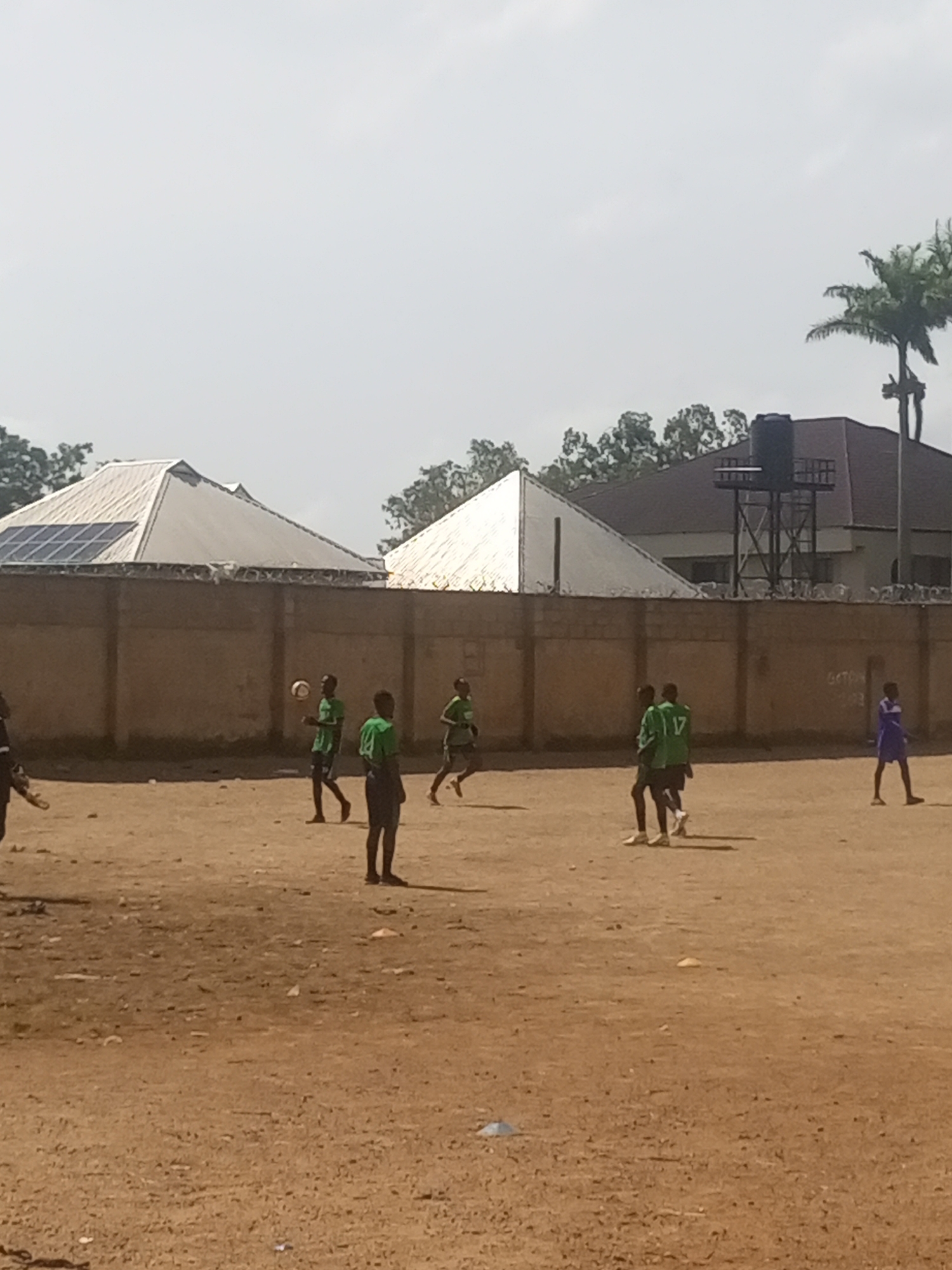 This image depicts an outdoor scene on a dirt field, likely a sports ground, under a bright sky. Several young males, appearing to be teenagers, are engaged in a soccer game. Most are wearing green jerseys and dark shorts, with one individual in a purple uniform. The players are scattered across the field, with some actively participating in the game and others observing. One player in the foreground is mid-stride, while another is positioned to receive a pass from a teammate. In the background, a low, tan-colored brick wall encloses the area. Beyond the wall, two buildings with distinctive metallic, angular roofs are visible. One of these buildings has solar panels installed on its roof. A water tank on a metal structure stands behind the other building, and a palm tree is visible to the right of the buildings. The ground is dry and sandy, with scattered debris. The overall impression is of a casual sports activity taking place in a somewhat undeveloped urban or semi-urban environment, potentially in Jos, Nigeria, given the architectural style and vegetation.