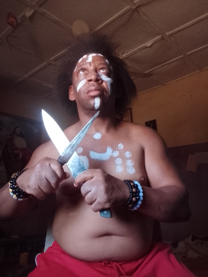 An adult male is observed indoors in Narayi, Nigeria. He possesses dark, voluminous hair and light-colored eyes directed upwards and to the right. White markings adorn his face and upper torso: a cross pattern on the forehead, a vertical line below the nose, horizontal lines on the cheekbones, a curved line on the left upper chest, and multiple circular patterns on the right chest and upper abdomen. He is shirtless and wears red shorts with a white trim. Multi-colored beaded bracelets are present on both wrists. He holds two knives in a crossed configuration before his chest: a knife with a wide, dull-silver blade and a black handle is in his right hand, angled downwards, while a narrower, pointed, dull-silver bladed knife with a dark handle is in his left hand, angled upwards. The blades exhibit signs of wear. The background features a light-colored, panel-style ceiling and a light-brown wall. A faded poster depicting a figure consistent with religious iconography of Jesus Christ is visible on the left wall, with partially discernible text "IL" beneath it. The scene is illuminated by ambient light.