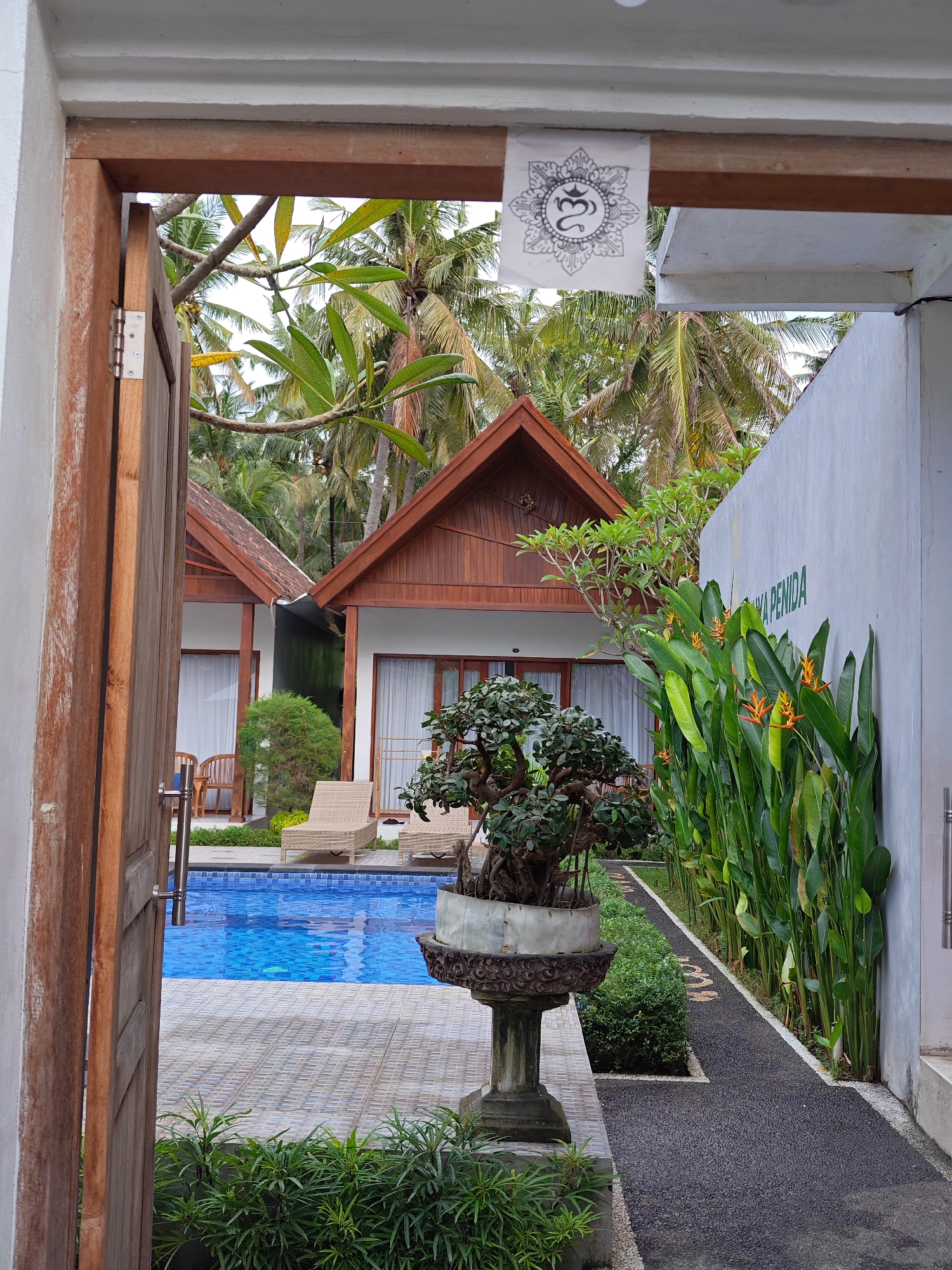 The image shows a view of a tropical resort from a doorway or entrance. The location is Nusa Penida, Indonesia, as indicated by the partial signage on the wall to the right which reads "...YA PENIDA." Through the doorway, two identical bungalows are visible, with brown wooden exteriors and peaked roofs, set back from a pool. The pool is blue and surrounded by tiled decking. Two lounge chairs sit on the decking in front of each bungalow. Palm trees and other tropical foliage fill the background. In the foreground, a bonsai tree in a decorative pot sits on a pedestal. A narrow walkway runs alongside a wall, lined with tall green plants with orange flowers. A small paper with a symbol printed on it is attached to the wooden beam of the doorway. The doorway itself is partially open, revealing the scene within. The scene suggests a calm and luxurious vacation setting.