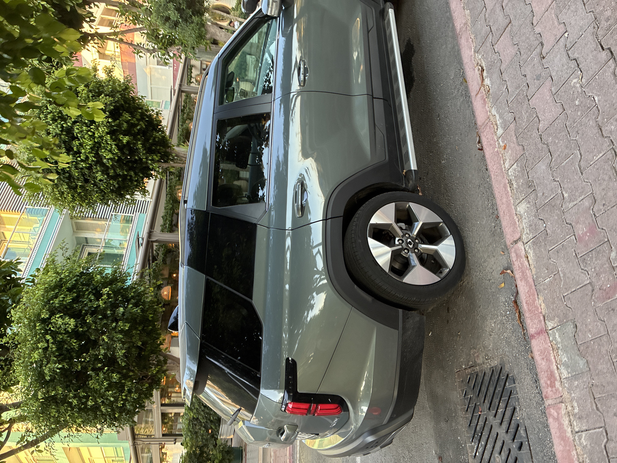 A grey SUV is parked on a city street in Alanya, Turkey. The vehicle is positioned on the right side of the road, facing away from the camera. The street is paved with asphalt and has a brick sidewalk on the right. A metal grate is visible on the street to the left of the vehicle. In the background, there are trees and a multi-story building with a light blue facade and balconies. The sun is casting shadows on the scene, suggesting it is late afternoon or early evening. The car has tinted windows and distinctive red taillights with a black accent.
