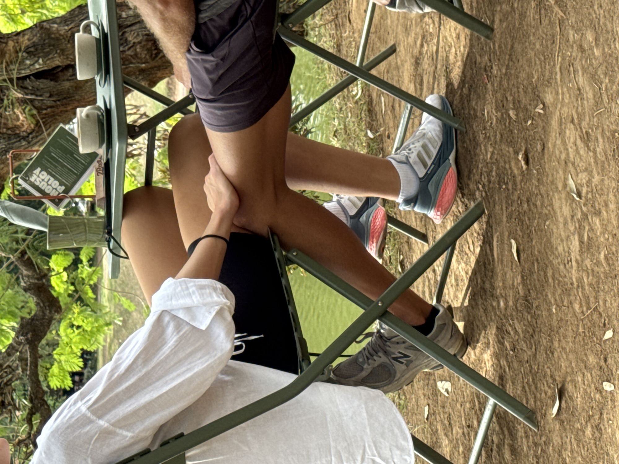 The image shows a close-up view of two people sitting at a green metal table, likely outdoors at a cafe or restaurant in Kuta Selatan, Indonesia. The person on the left is wearing a white long-sleeved shirt and black shorts, with a black bracelet on their left wrist. Their left foot is resting on the ground, wearing a grey New Balance sneaker. The person on the right is wearing grey shorts and white Adidas sneakers with pink accents on the soles. Their legs are crossed at the knees, and their right hand rests on the left person's leg. On the table are two cups and saucers, a green cup, and a sign with "HOUSE POLICY" printed on it. The background features lush green foliage and the trunk of a large tree, suggesting a natural, possibly tropical setting. The ground appears to be dirt or dry leaves, with some scattered leaves. The lighting indicates it is daytime.