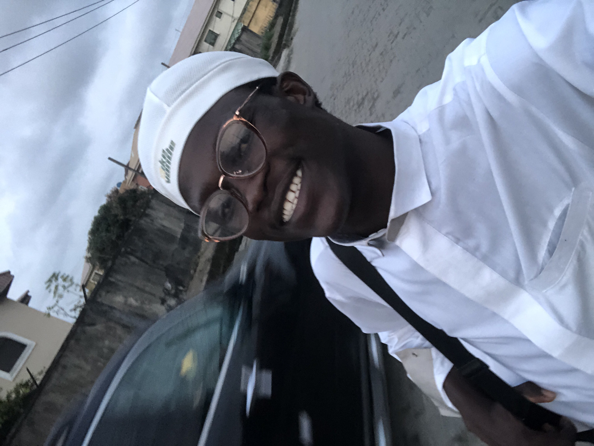 In Ado, Nigeria, an over-the-shoulder selfie captures a young Black man smiling. He wears a white kufi cap and a white collared shirt. His round, translucent glasses have a slight sheen, reflecting light. A dark strap from a bag crosses his chest. Behind him, a dark vehicle's tinted window is visible, along with a concrete wall and a building with a pitched roof. The ground appears to be paved, and the sky is overcast.
