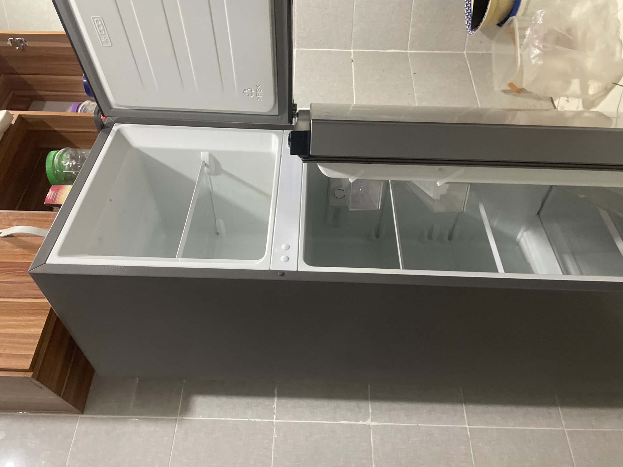The image shows an open chest freezer, with the lid raised. The freezer has two compartments. One compartment is visible and appears empty, separated by a white divider. The other compartment has three dividers, but the contents are not visible. The freezer is dark gray.
To the left of the freezer is a wooden cabinet. The freezer is situated on a tiled floor.
The location of the image is unknown, and therefore, cannot be determined to be Abuja, Nigeria.
