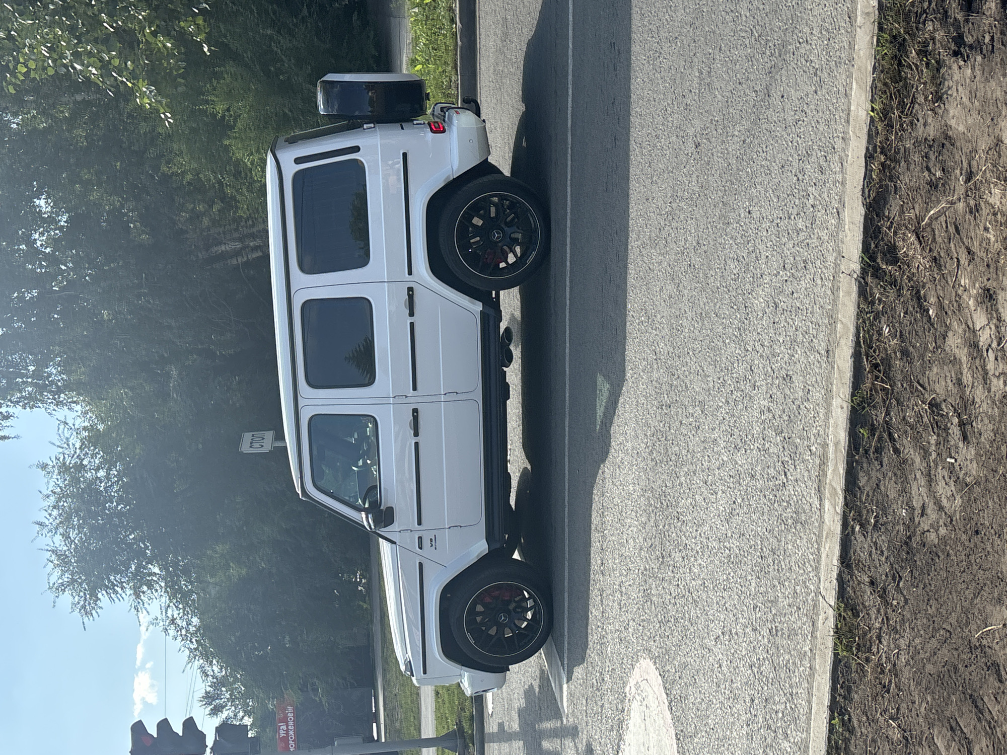 A white Mercedes-Benz G-Class SUV is parked on the right side of a paved road in Novosibirsk, Russia. The vehicle is facing right, with its front wheels turned slightly towards the left. The car has black rims, red brake calipers, and a spare tire mounted on the rear. A red traffic light is visible on the left side of the image, along with a sign that reads "СТОП" (STOP in Russian). The background consists of lush green trees and a bright blue sky. The ground in the foreground is a mix of asphalt, dirt, and sparse vegetation.