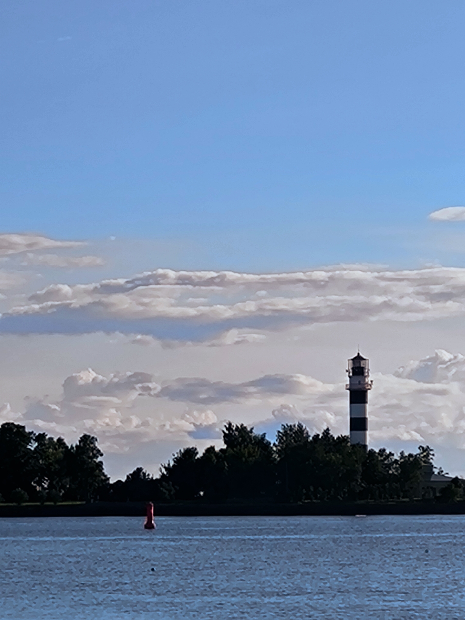 The image depicts a serene waterscape under a bright, cloudy sky. In the foreground, a calm body of blue water stretches across the frame, with gentle ripples suggesting a light breeze. A single, bright red buoy floats in the water towards the left side of the image, marking a point of interest or a navigational hazard.
In the middle ground, a dark, dense line of trees forms the coastline, creating a rich green backdrop. Rising above the trees, towards the right side of the image, stands a striking lighthouse. This lighthouse is characterized by its black and white striped pattern, a classic design that stands out against the natural surroundings and the sky. The top of the lighthouse features a lantern room with glass panels and a pointed roof.
The sky above is a vibrant blue, punctuated by numerous clouds. These clouds vary in texture and color, with some appearing bright white and fluffy, while others are more diffused and tinged with grey, hinting at varying atmospheric conditions. The lighting suggests it is daytime, likely late afternoon given the soft quality of the light and the contrast between light and shadow.
The overall mood is peaceful and picturesque. The scene evokes a sense of calm and highlights the interplay between the natural environment and man-made structures. The location is Riga, Latvia, and while no specific landmarks are explicitly visible other than the lighthouse, the scene is consistent with coastal or riverine scenery found in such a region. There is no visible text in the image.