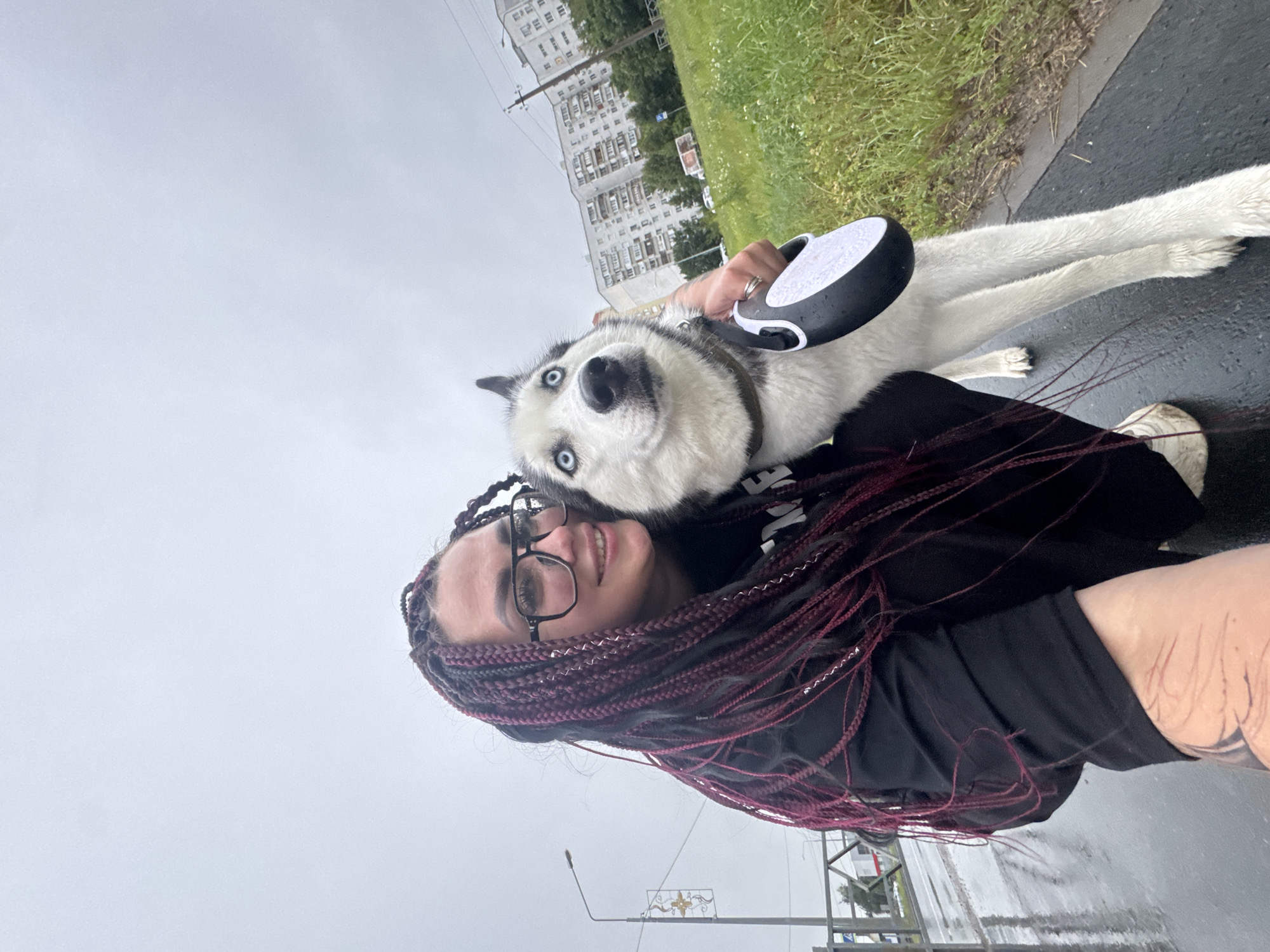 A person with dark skin, eyeglasses, and long, braided, burgundy-colored hair is taking a selfie outdoors with a husky dog. The person is wearing a black hoodie and white sneakers. The husky has striking blue eyes and is wearing a collar. The person's right hand is holding a black and white retractable dog leash. They are standing on a wet asphalt surface, likely a sidewalk or path, with a grassy area and buildings visible in the background. The sky is overcast. The person's left arm, visible in the foreground, has a tattoo on it. The scene appears to be in a residential area of Tolyatti, Russia, indicated by the style of the apartment buildings.
