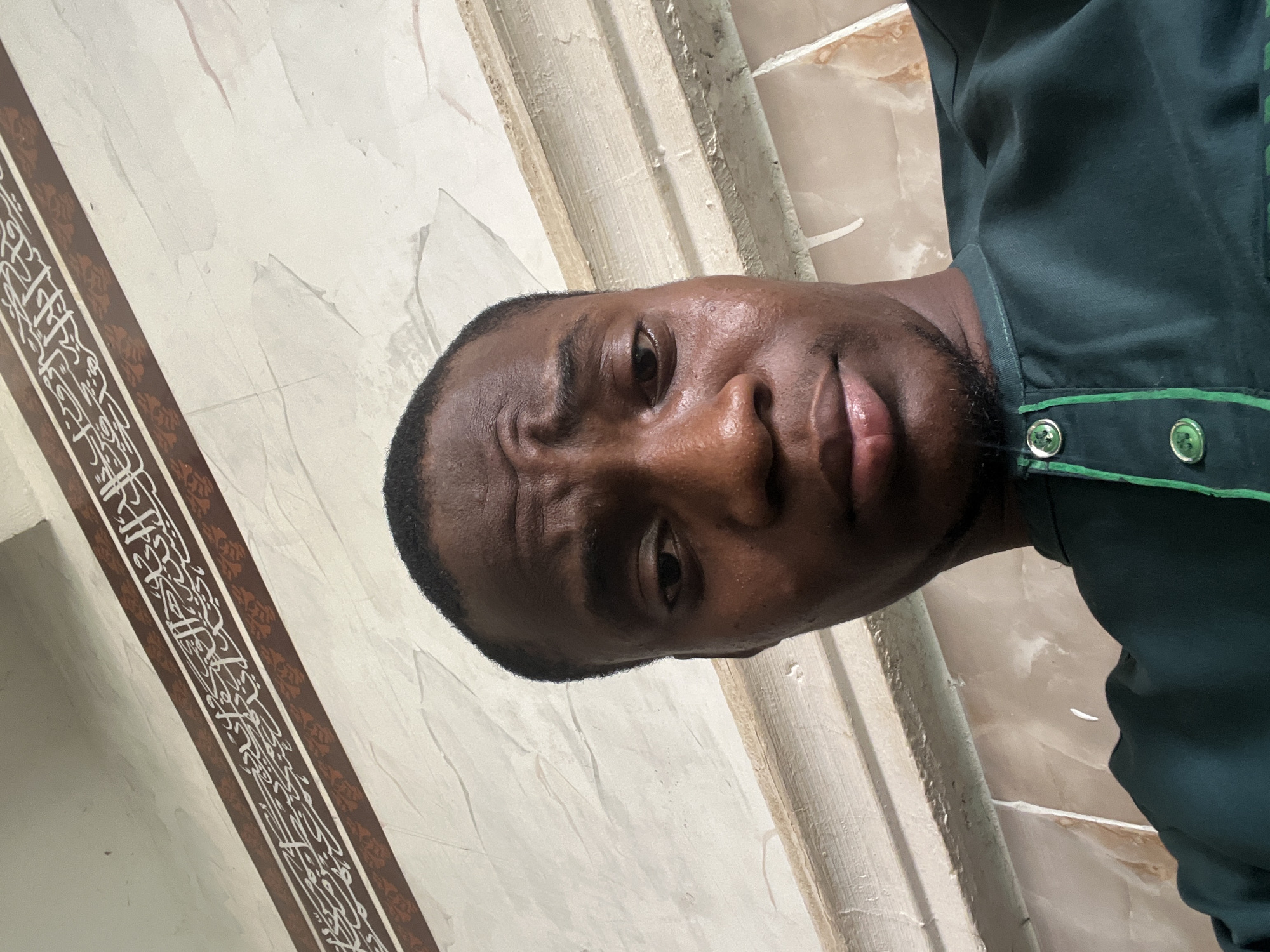 A selfie-style image shows a dark-skinned man with a close-cropped haircut. He is looking directly into the camera with a slight frown. He wears a dark green shirt with a light green trim and two visible silver buttons. The background is a wall with a light beige and white marble-like pattern. A decorative band with brown borders and Arabic calligraphy is visible on the left side of the image. The location is in Katcha, Nigeria.