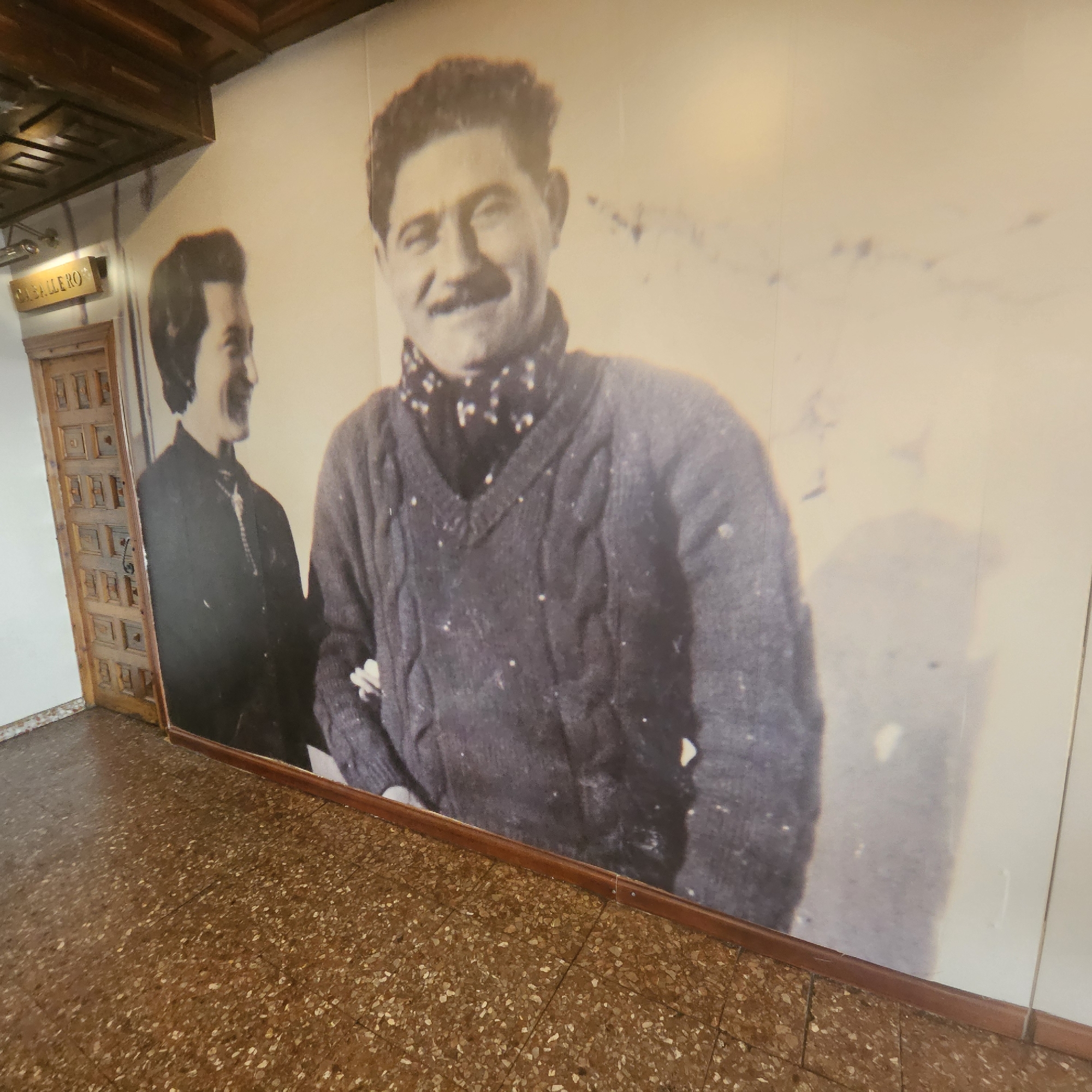 The image shows a mural of two men on a wall, with a wooden door and a patterned floor in the foreground. The mural appears to be in a public space, possibly a museum or a historical building, located in Almadrones, Spain. The man on the right is a close-up, showing his face and upper body. He has dark, curly hair, a mustache, and is wearing a dark V-neck sweater with a patterned scarf around his neck. The man on the left is partially visible, standing next to the other man, wearing a dark jacket and a scarf. A sign with the word "CABALLERO" is visible to the left of the mural, above the wooden door. The floor is covered with small, multi-colored stones, typical of Spanish terazzo flooring. The overall scene depicts a historical or cultural display.