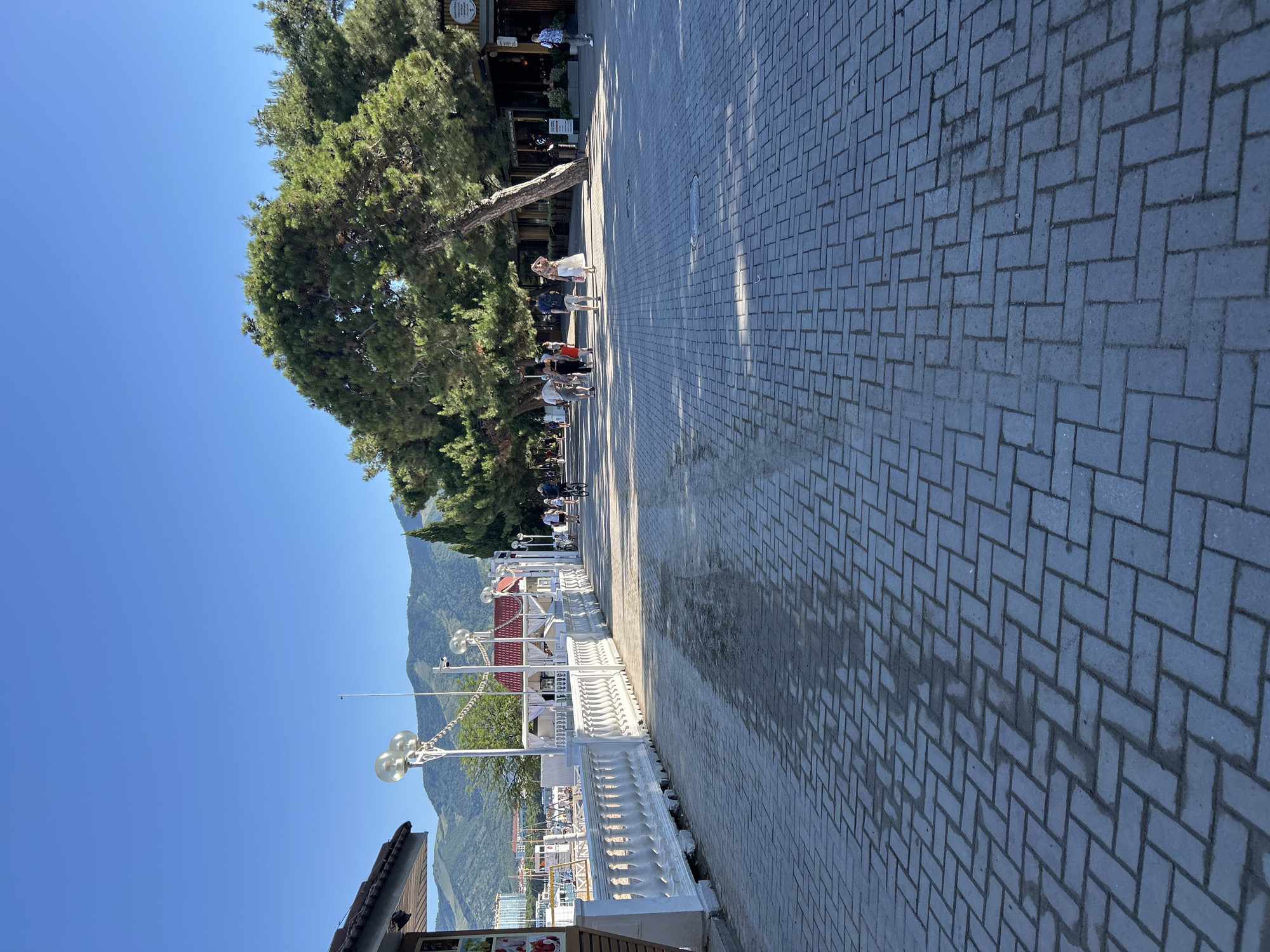 The image shows a paved promenade in Gelendzhik, Russia, on a clear, sunny day. The promenade is lined with white ornate railings and streetlights on the left, with buildings and trees on the right. Several people are walking and cycling along the promenade. In the background, there are mountains and a clear blue sky. The scene depicts a lively public space, likely a tourist area.