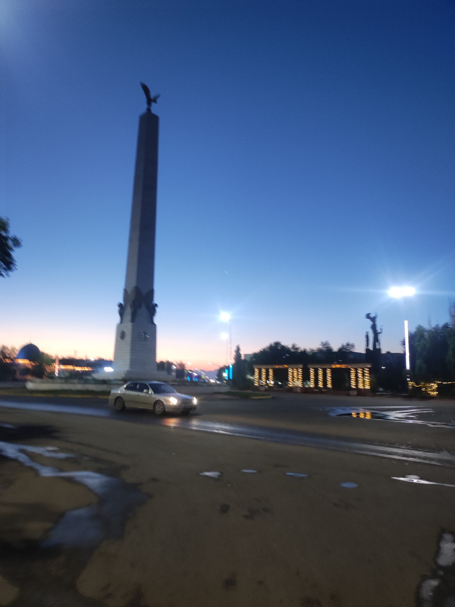 The image depicts a street scene in Hromtau, Kazakhstan, during twilight. A tall, slender obelisk, topped with a bird silhouette, dominates the left side of the frame. The obelisk appears to be a monument. A silver sedan is captured in motion, driving on a wet asphalt road in the foreground. To the right of the obelisk, a dark silhouette of a statue or monument stands against the twilight sky, illuminated by streetlights. Decorative lights are strung across an archway or structure in the background, creating a festive atmosphere. The sky transitions from a deep blue at the top to a lighter hue near the horizon, hinting at the fading light. The road shows reflections of the lights and what appear to be puddles, suggesting recent rain. Trees are visible in the middle ground, contributing to the urban landscape.