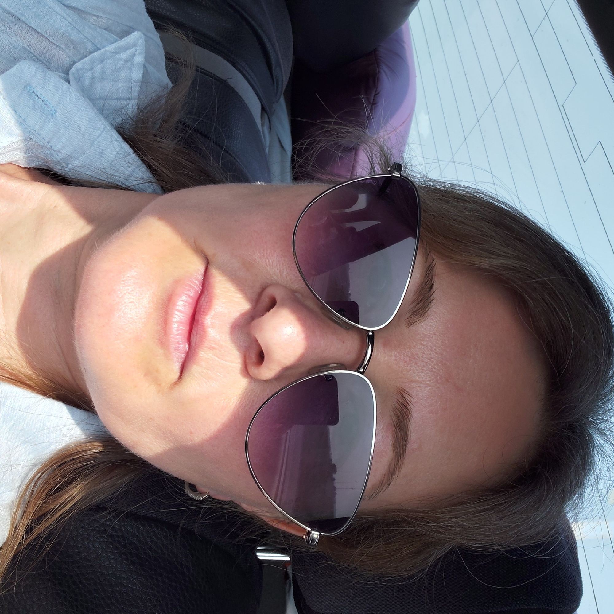 A woman is taking a selfie in a car. She is wearing sunglasses and a light blue collared shirt. The background shows the interior of the car, including a headrest and a window with a sunshade. A purple pillow is visible in the back seat. The lighting suggests it is daytime. The image appears to be taken inside the city of Ubinskoye, Russia.