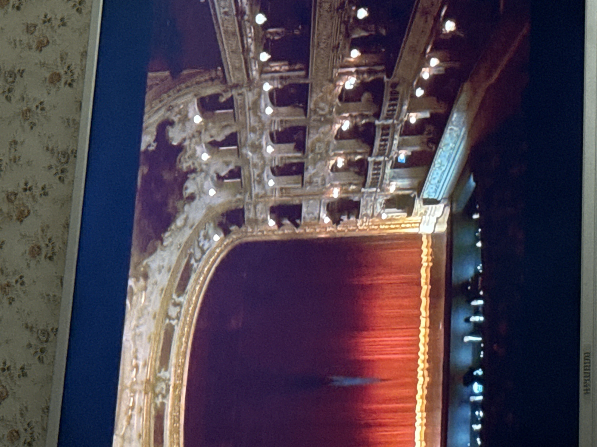 The image shows the interior of an ornate theater, featuring a stage with a red curtain, a ceiling with decorative boxes and lights, and detailed architectural ornamentation. The image is a photograph of a screen displaying this theater scene, with a section of floral wallpaper visible on the left side. The theater is likely not located in Mytishchi, Russia.