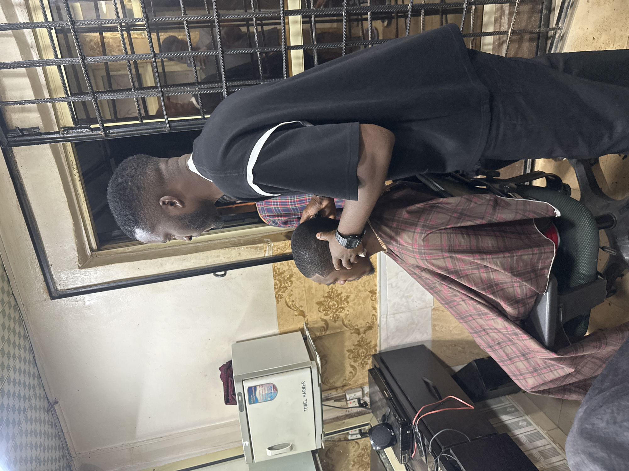 An indoor scene within a barber shop or hair salon depicts two men. On the right, a standing Black male, identified as the barber, with a short haircut and beard, wears a black short-sleeved t-shirt with white stripes and black trousers. He is positioned behind and to the left of a seated Black male client, whose head is slightly tilted forward. The barber's right hand is near the client's head, suggesting an ongoing haircut or styling, and he wears a black digital watch on his left wrist. The client is draped in a brown and pink plaid barber cape.
To the left of the client, a white "TOWEL WARMER" appliance, bearing an "ELECTRIC TOWEL WARMER" sticker, is mounted on the wall, with a dark red cloth resting on top. Below it, a dark-colored counter holds electronic equipment including an LG-branded speaker and another device, connected by red and black wires. A mirror behind the counter reflects parts of the wall and electrical outlets. The lower section of the walls features a golden-brown ornate patterned wallpaper, while the upper section is plain cream. The ceiling is a grid of light-colored patterned panels. On the right side of the image, behind the barber, a doorway or window opening is secured with black metal bars, through which indistinct reflections of figures and an indoor environment are visible. The floor appears to be tiled in a light hue. The general setting is a functional barber shop in Unknown, Unknown.