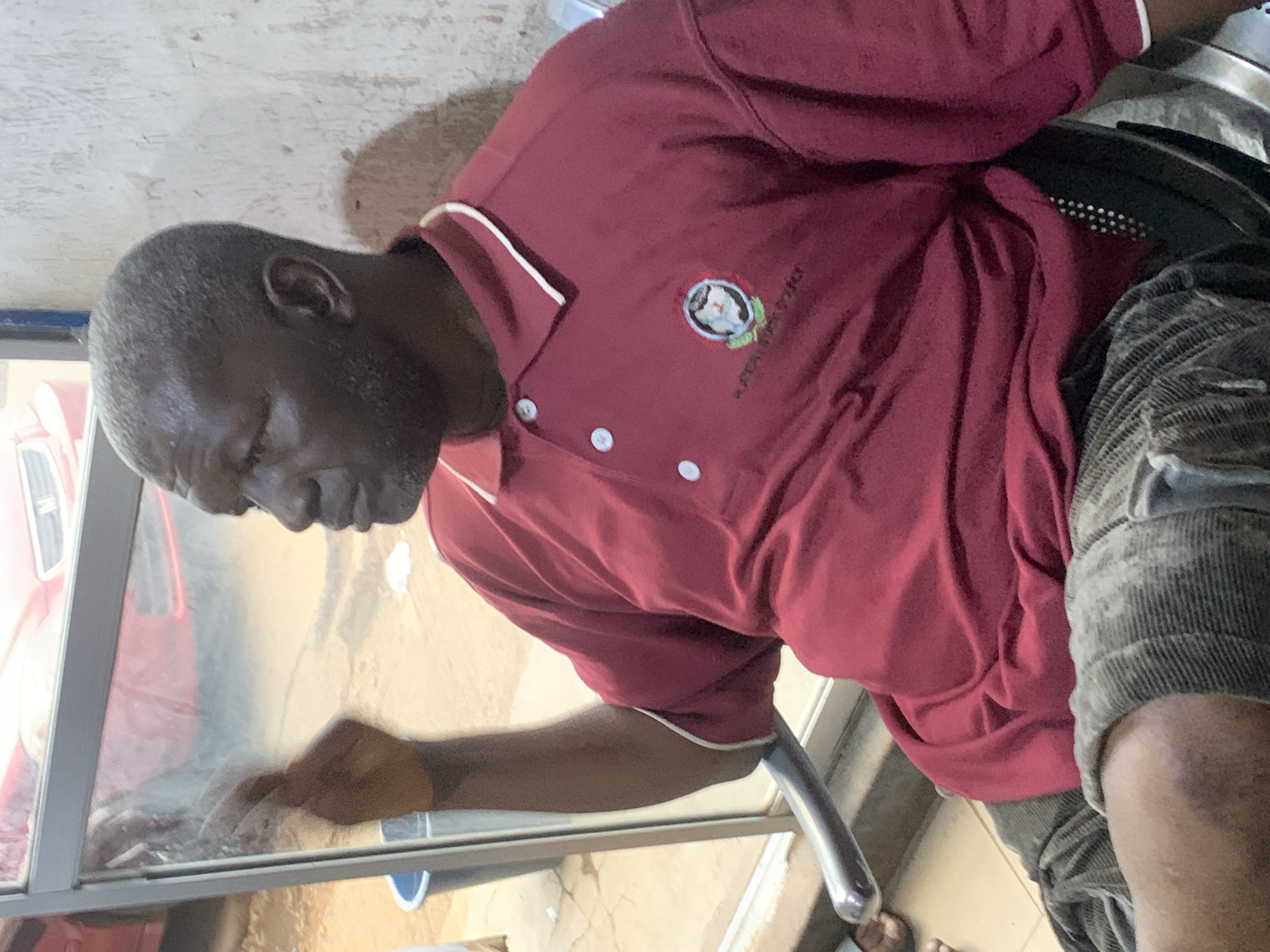 An older Black man in a maroon polo shirt is wiping a glass surface. The shirt has white trim and a small emblem on the chest. He wears gray textured shorts and appears to be seated. The scene suggests an indoor environment, possibly a commercial space. The location is implied to be Jos, Nigeria.