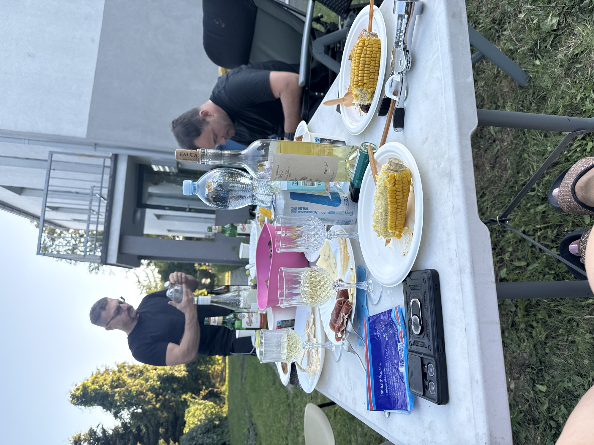 A group of people are having an outdoor gathering at a table laden with food and drinks. The table is set with plates of grilled corn on the cob, slices of meat, cheese, and glasses of white wine. Several bottles of wine and beer, along with a large bottle of water, are also on the table. A pink bowl sits in the center of the table. In the background, a man in a black t-shirt is seen pouring a drink. Another man is seated in a chair, looking down at the table. The gathering appears to be taking place in a backyard with lush green grass and trees. The setting is consistent with a private residence, possibly in Katowice, Poland, given the context.
