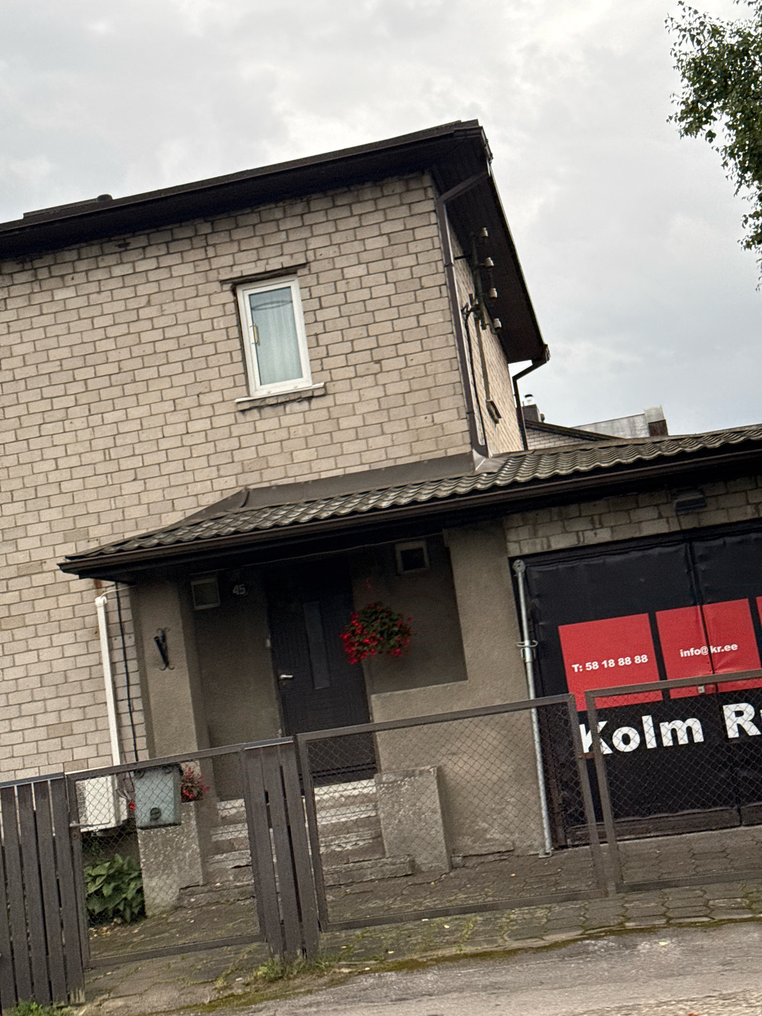 This is an exterior, eye-level, slightly tilted, medium shot of a brick building in Tallinn, Estonia. The building appears to be a residential house with a garage attached. The main house is made of light beige bricks with a dark brown gutter and roof. A single, small white-framed window is visible on the upper floor. The attached garage has a black roll-up door with red lettering on it. The lettering on the door says "T: 58 18 88 88" and "info@kr.ee", and "Kolm R". In front of the garage is a dark metal fence. To the left of the garage, there is a dark-colored door leading into the house, with a hanging basket of red flowers above it. Next to the door is a small electrical box. The ground in front of the building is paved with cobblestones. A tree with green leaves is visible on the right side of the frame. The sky is overcast and grey.