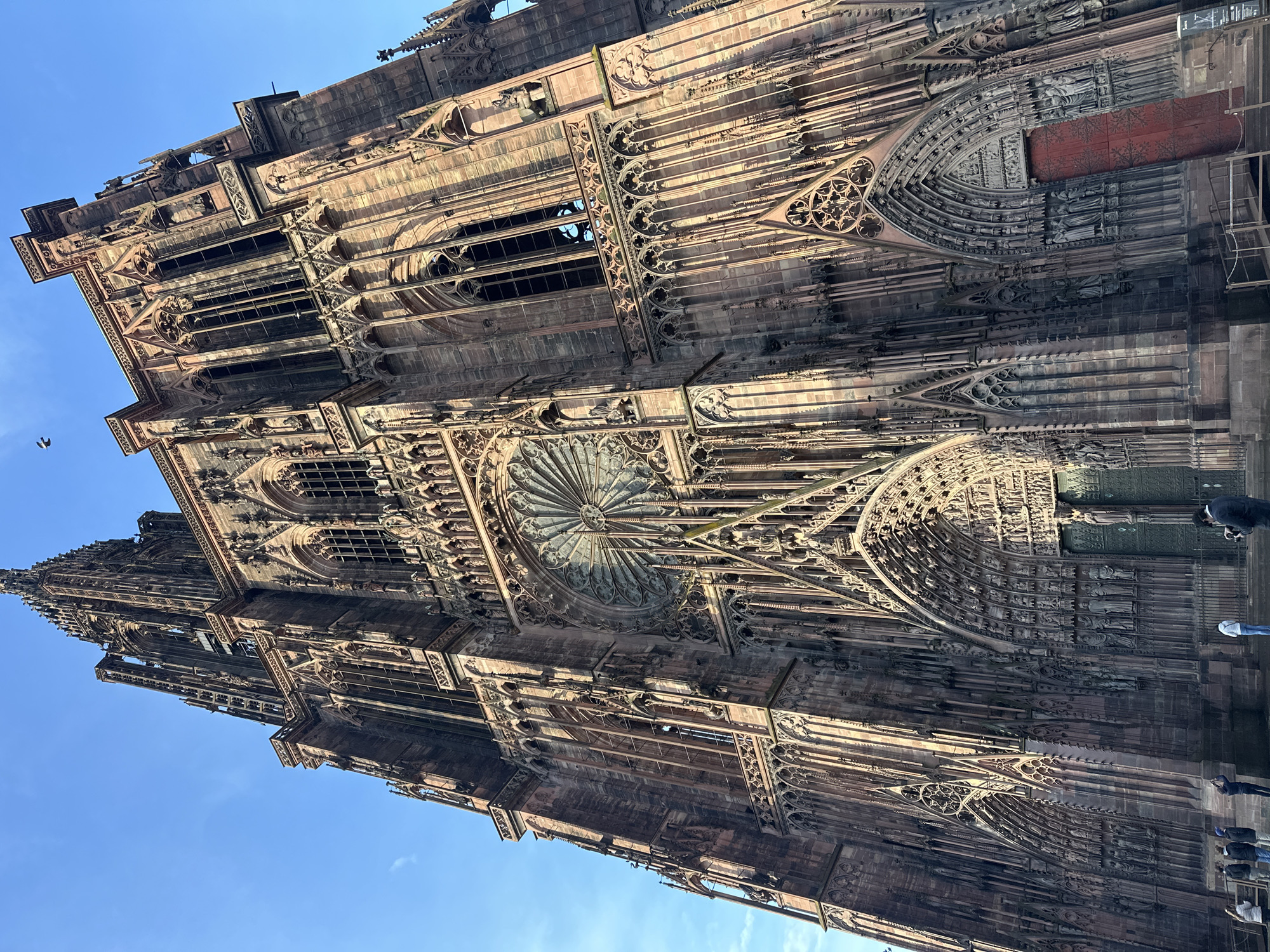 The image shows a low-angle, upward view of the facade of Strasbourg Cathedral (Cathédrale Notre-Dame de Strasbourg) in Strasbourg, France. The sandstone building exhibits intricate Gothic architectural details, including elaborate carvings, a rose window, spires, and pinnacles. The facade is composed of multiple levels with different architectural features. A blue sky is visible in the background. Several people are standing near the base of the cathedral, partially visible in the lower-right of the frame.