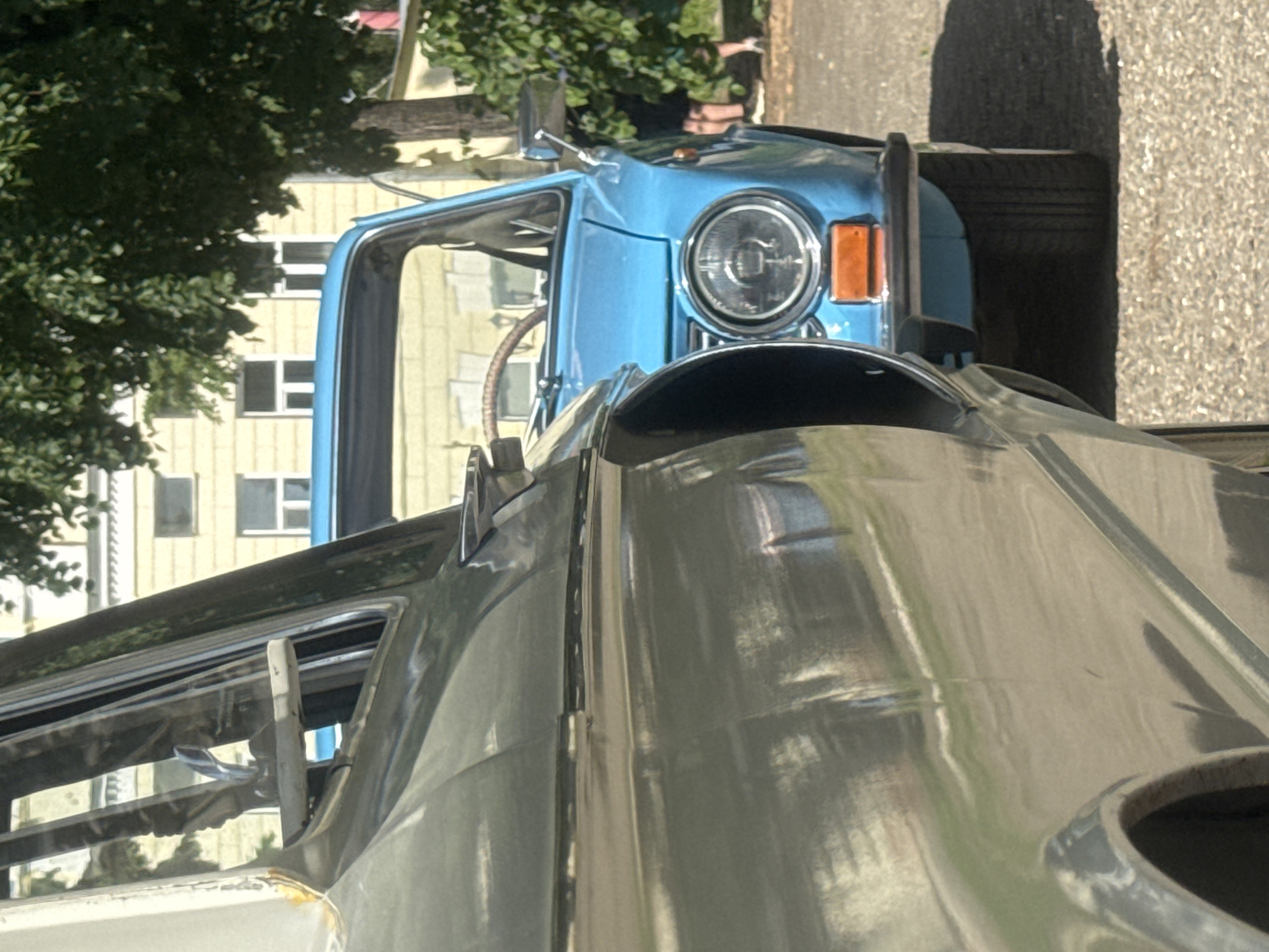 The image depicts the front of two vehicles parked on a paved surface. The vehicle in the foreground is a silver-colored car with a prominent hood and windshield. The vehicle behind it is a light blue car with a round headlight and orange signal light.
In the background, a building with white-framed windows is visible behind a tree. The location is possibly Kazan, Russia, based on the presence of Soviet-era vehicles and building architecture, though this cannot be confirmed without further information.
No people are clearly visible in the image. The focus is on the cars and the surrounding environment.