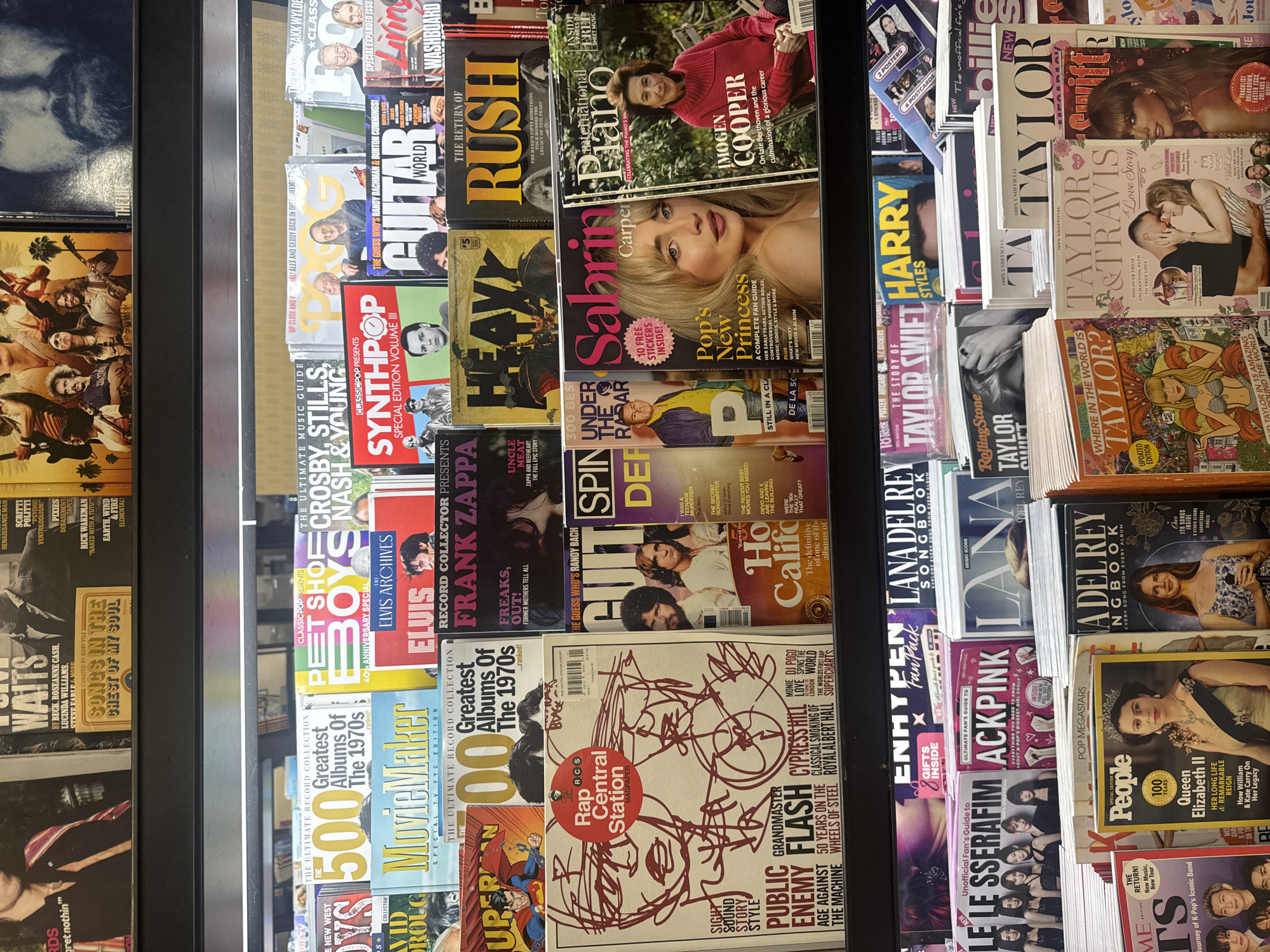 The image depicts a multi-tiered retail magazine display rack in Boca Raton, United States of America, presenting an array of music, entertainment, and celebrity-focused periodicals. The magazines are arranged across four visible shelves, illuminated by overhead fluorescent lighting.
On the top visible shelf, partially obscured, titles include "SONGS IN THE CHEST OF MY SOUL" and "EARTH, WIND & FIRE."
The second shelf features numerous magazines. Prominent titles include "THE 500 Greatest Albums Of The 1970s," "PET SHOP BOYS 40TH ANNIVERSARY SPECIAL," "CROSBY STILLS NASH & YOUNG," and "SYNTHPOP SPECIAL EDITION VOLUME III" displaying multiple individuals. Other titles include "GUITAR WORLD," "WASHBOARD," and "ELVIS ARCHIVES" with a small image of Elvis Presley.
The third shelf showcases "MovieMaker SPECIAL TV FYC EDITION," a "SUPERMAN" comic cover, and "Greatest Albums Of The 1970s." Further along are "FRANK ZAPPA" with text "FREAKS, OUT! FORMER MOTHERS TELL ALL," "HEAVY," and "THE RETURN OF RUSH." A large "Rap Central Station MagPaper" features red graffiti-style line art of faces and lists "PUBLIC ENEMY," "GRANDMASTER FLASH," and "CYPRESS HILL." Next are "SPIN," "UNDER THE RADAR" featuring a man in a yellow jacket, "Sabrina Carpenter" with a portrait of a young blonde woman, "International Piano" featuring an older woman in a red sweater, and "Ho Califo."
The fourth, lowest visible shelf contains magazines such as "ENHYPEN FanPack," "LE SSERAFIM" featuring five women, "ACKPINK" about a K-pop group, and "LANA DEL REY SONGBOOK." At the very bottom, "TIME" with "THE RETURN! New Music, New Tour" is visible. A "People" magazine cover displays a portrait of Queen Elizabeth II. Another "A DEL REY SONGBOOK" features Lana Del Rey. Other prominent covers include "WHERE IN THE WORLD IS TAYLOR?" in a graphic novel style, and "TAYLOR & TRAVIS A Love Story" depicting Taylor Swift and Travis Kelce embracing. Multiple other magazines focused on "TAYLOR SWIFT" are also present.