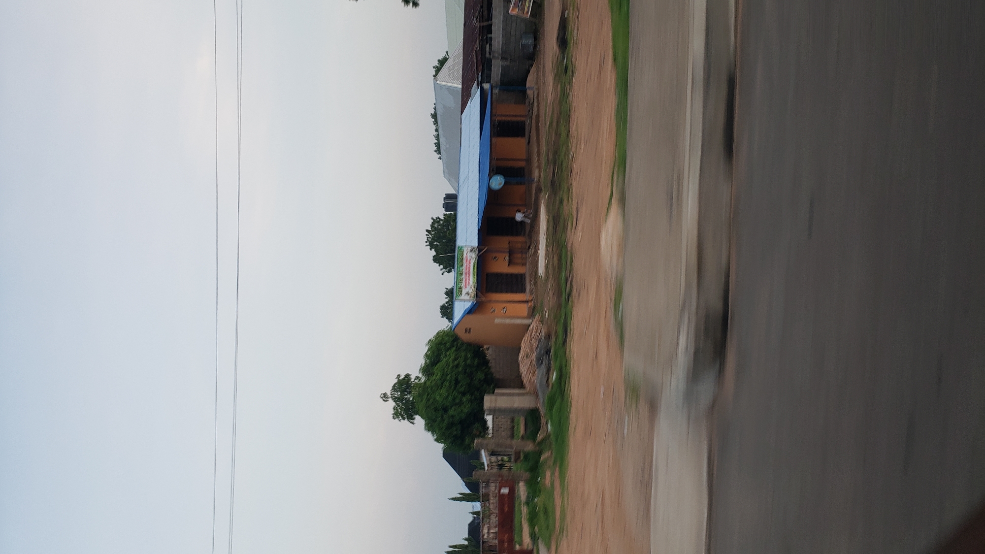 This image, taken from a moving vehicle on a road in Jalingo, Nigeria, captures a roadside scene. In the mid-ground, a single-story building with an orange facade and a blue corrugated metal roof stands prominently. The building appears to house a business, as indicated by a green sign with white text above the entrance, which is partially obscured. Adjacent to the building, piles of what seem to be construction materials, possibly sand and bricks, are visible. A lush green tree is situated to the left of the building, casting a shadow. In the background, other structures and trees can be seen, suggesting a semi-urban or developed rural area. The foreground is dominated by a blurred road and a concrete median, indicating motion. The sky above is pale and overcast.