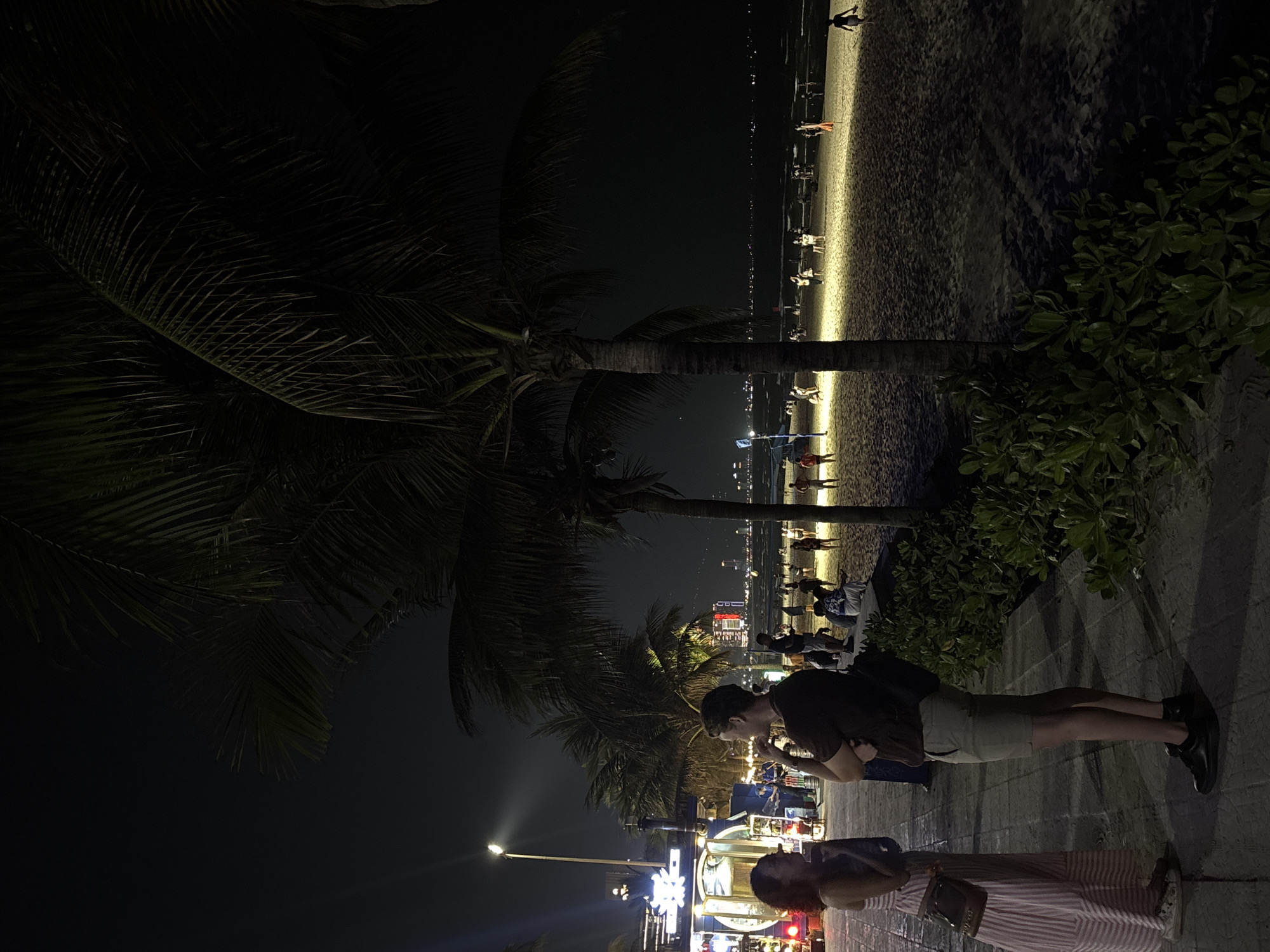 A vertical outdoor shot shows a nighttime scene at a beach in Đà Nẵng, Vietnam. A couple stands on a tiled walkway facing the beach, partially obstructing a row of brightly lit businesses. Behind them, palm trees frame a dimly lit beach with several people walking along the water's edge. The background reveals city lights along the shoreline.
