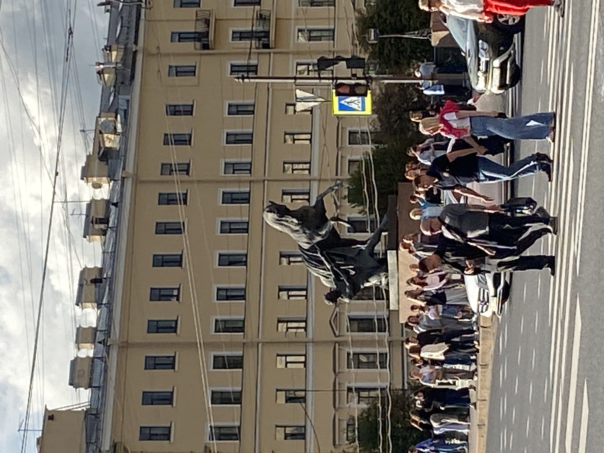 The image depicts a street scene in Saint Petersburg, Russia, likely in front of an apartment building with a pale yellow facade and multiple windows. A large bronze equestrian statue dominates the center of the frame, featuring a man on a rearing horse. A group of pedestrians is gathered on the sidewalk behind a barrier, observing the statue. In the foreground, several people are crossing a street at a pedestrian crossing. Traffic lights and street signs are visible, indicating an urban environment. The sky is partly cloudy, suggesting daytime.