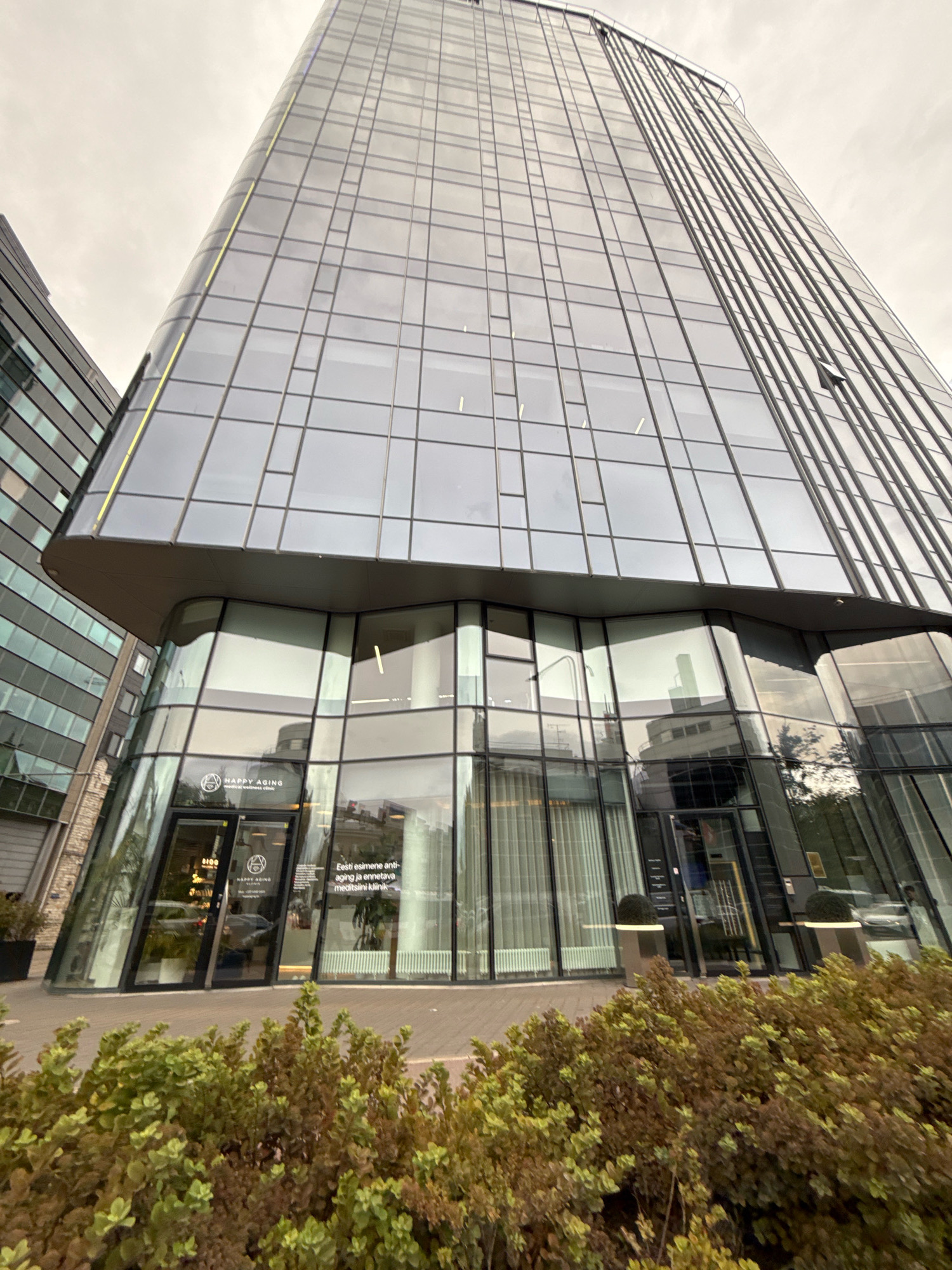 The image shows the exterior of a modern, glass-fronted building in Tallinn, Estonia. The building is a skyscraper with a curved facade covered in reflective glass panels. In the foreground, there are bushes and low-lying plants. The entrance to the building is visible at the base of the skyscraper, with glass doors and signage that reads "HAPPY AGING medical & wellness clinic" in English, and "Eesti esimene anti-aging ja ennetava meditsiini kliinik" in Estonian. The sky is overcast, suggesting a cloudy day. The overall impression is one of a contemporary urban setting.