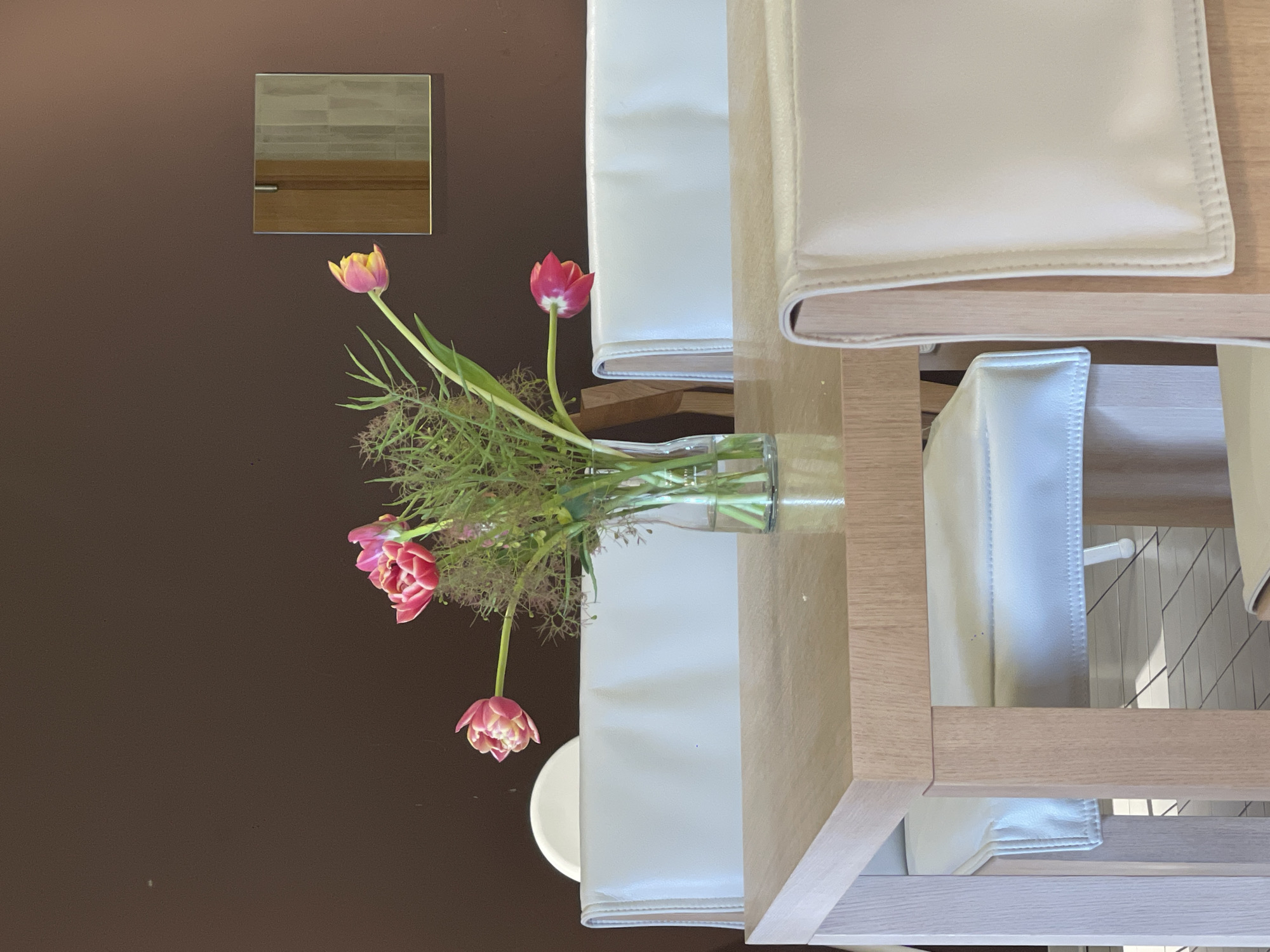 A medium shot taken indoors shows a vase with tulips and greenery on a light-colored wooden table. The vase is clear glass and the tulips are a mix of red and pink with some yellow. The greenery consists of various types of leaves and sprigs. Behind the vase, there are two white cushioned chairs stacked on top of each other, with light wood frames. On the brown wall to the left, there is a small square mirror, and beneath the vase, there is a white plate. The setting does not allow for the identification of the specific location or city.