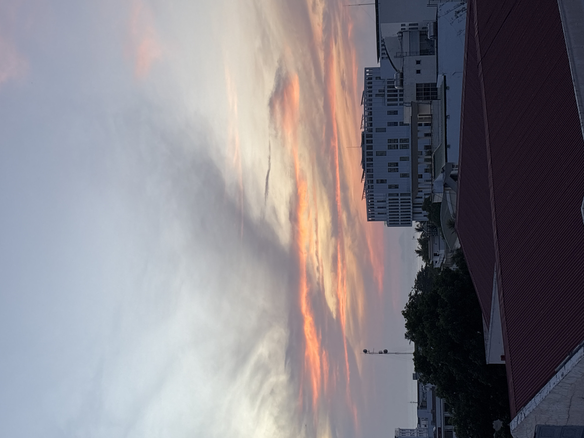 A rooftop view captures a city skyline at sunset. The sky is a canvas of soft blues, grays, and vibrant oranges and pinks, with wispy clouds stretching across the horizon. Dominating the mid-ground is a modern, white building with a distinctive rounded facade and numerous windows. In the foreground, two rooftops are visible, one with a dark red corrugated metal roof and the other with a lighter-colored, possibly stone, facade. A tall, slender tower with multiple antennas stands to the left of the central building. Trees with dark green foliage are scattered throughout the scene, interspersed with other buildings and structures. The ocean is visible in the distance, a hazy line between the city and the sky. The image appears to have been taken in Nha Trang, Vietnam, given the architectural style of the buildings and the coastal setting. No people are discernible in the image.