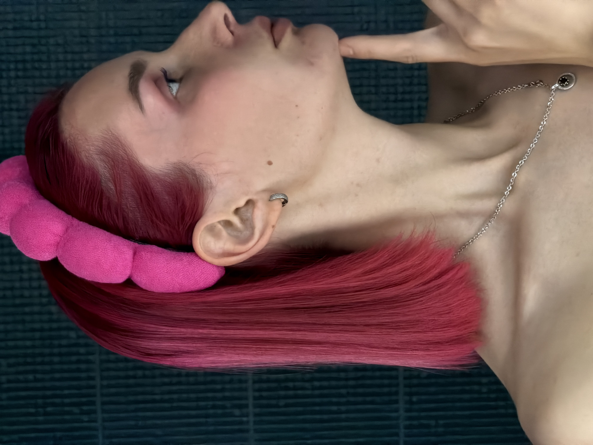 A profile view of a young woman with bright pink hair is shown against a dark green tiled background. She is wearing a pink plush headband and a silver necklace with a dark pendant. Her right hand is touching her chin, and she appears to be looking to the right. The image focuses on her face, neck, and shoulder. There are no discernible landmarks or elements that would indicate a specific location within the city of Chaikovsky, Russia.