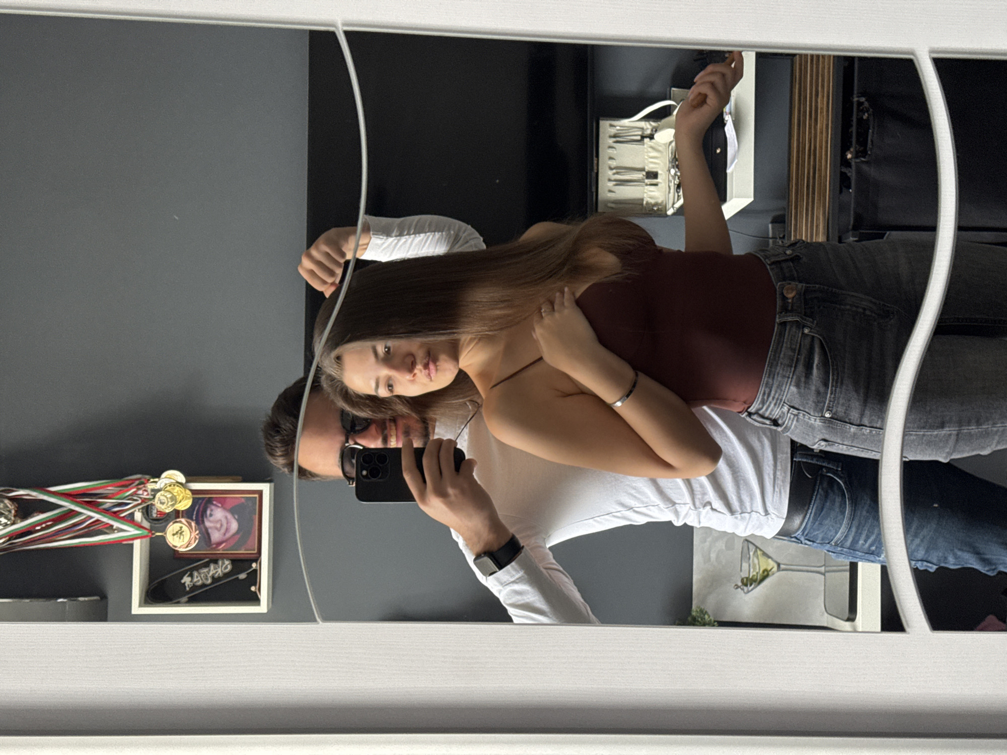A mirror selfie shows a couple. The man is holding a phone to take the picture, and the woman is standing beside him, her arm around his waist. The man is wearing sunglasses, a black watch, and a white long-sleeved shirt. The woman has long brown hair and is wearing a red tank top. In the background, there is a gray wall, a black wall, and a jewelry box. On a shelf, there is a framed picture, some medals, and a miniature skateboard. Below, there's an image of a martini glass with olives. The location is suggested to be Sofia, Bulgaria.
