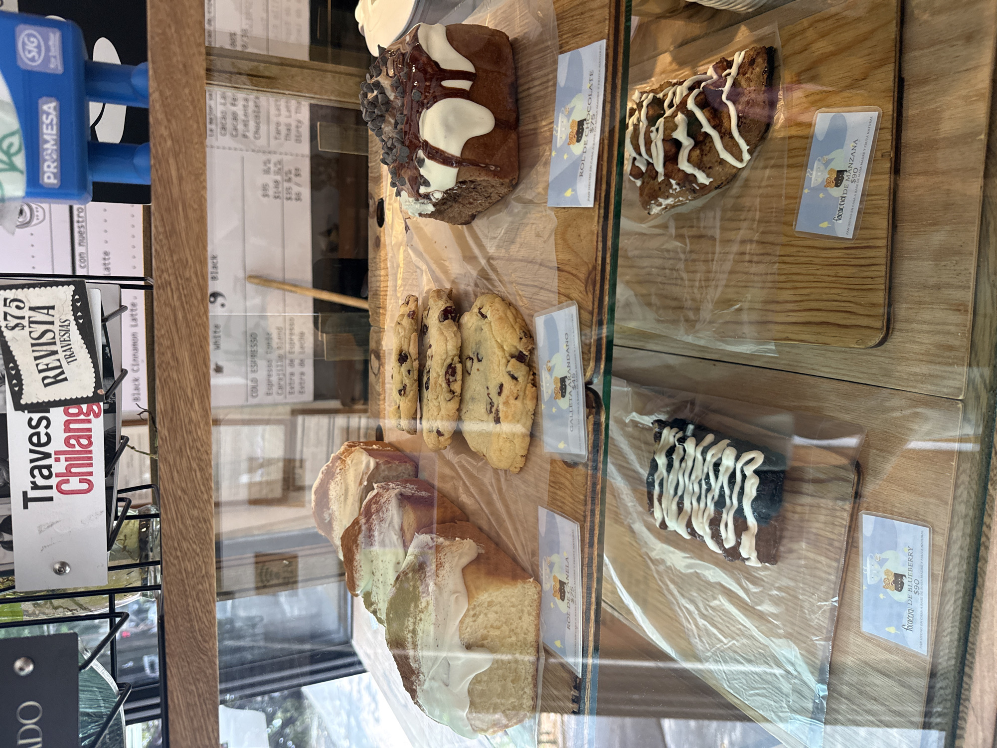 A bakery display case exhibits several types of pastries, including rolls, cookies, and focaccia, each in a plastic wrapper with a descriptive tag indicating the type and price. A "Rol de Canela" is $75, "Galleta Arandano" $45, "Rol de Chocolate" is on display with chocolate chips, "Focaccia de Blueberry" is $90, and "Focaccia de Manzana" is also $90. The display case also includes signage and a promotional figure. The location is likely in Mexico City, Mexico, as indicated by the "Travesias Chilang" tag.
