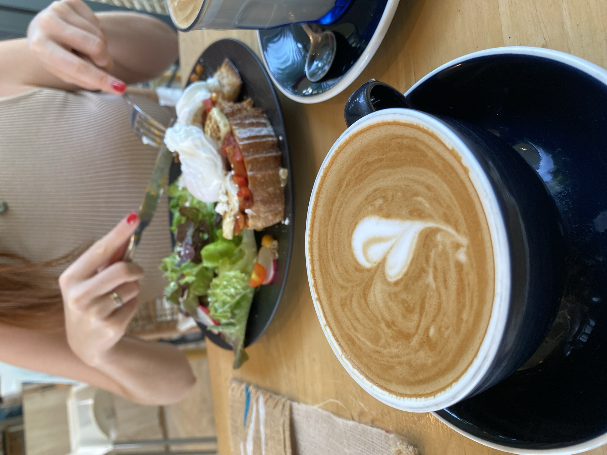 An overhead shot shows a woman seated at a wooden table in Choeng Thale, Thailand. She is holding a fork and knife, preparing to eat a dish featuring bread, poached eggs, and salad. To the right of the plate is a large, blue coffee cup with latte art. Above the plate, another glass cup sits on a saucer. A small burlap napkin rests on the table near the coffee cup. The focus is on the food and drink arrangement.