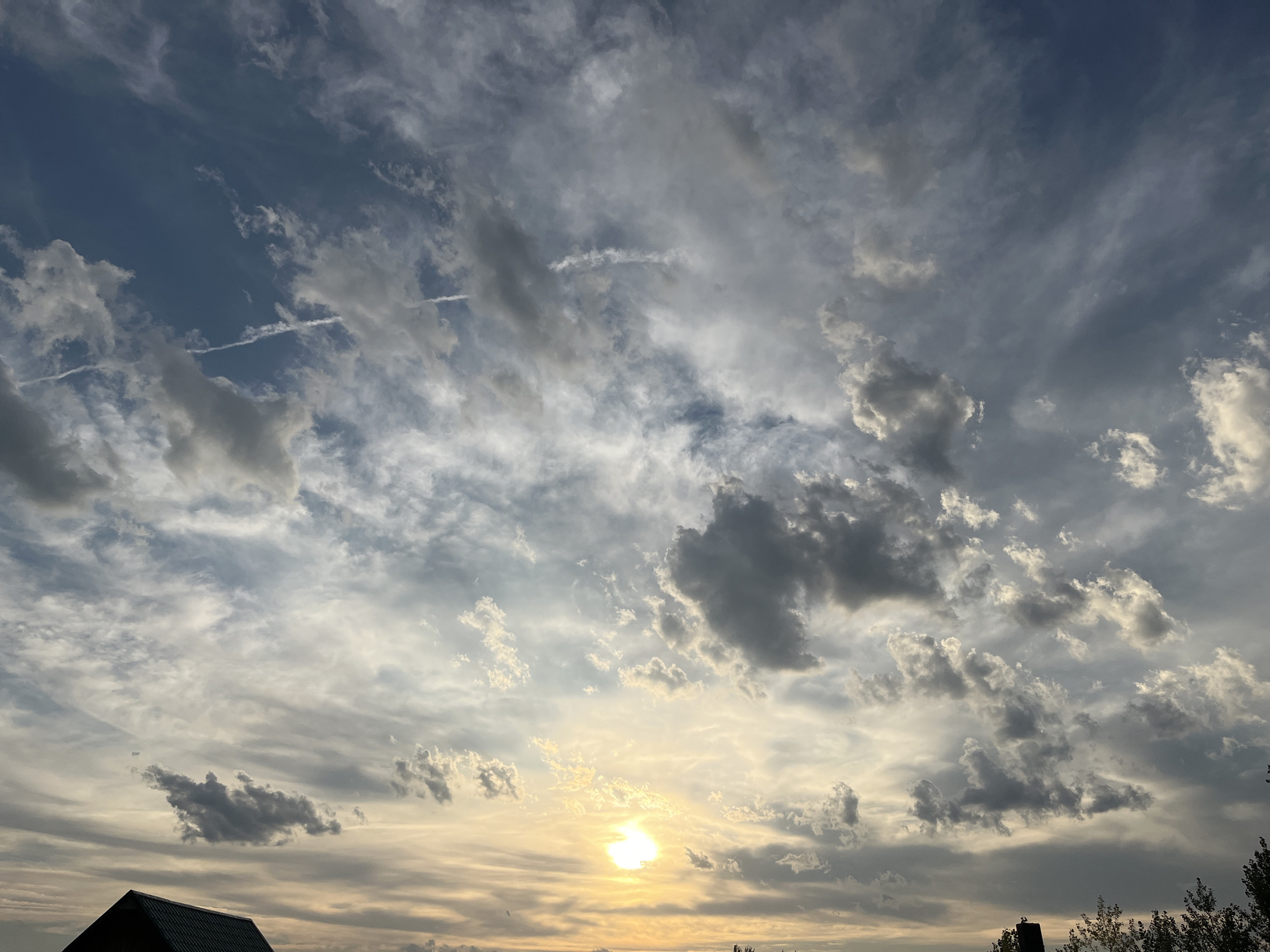 The image captures a dramatic sky over the city of Shumeyka, Russia, likely during sunset. The sky is filled with a mix of dark and light clouds, with a bright, hazy sun peeking through the lower center of the frame, casting a warm, golden glow. Wispy cirrus clouds stretch across the upper portion of the sky, interspersed with thicker, more textured cumulus clouds. A few faint contrails are visible, indicating air traffic. In the foreground, the dark silhouettes of the rooftops of buildings and trees are visible against the vibrant sky. The overall impression is one of a dynamic and atmospheric celestial display.