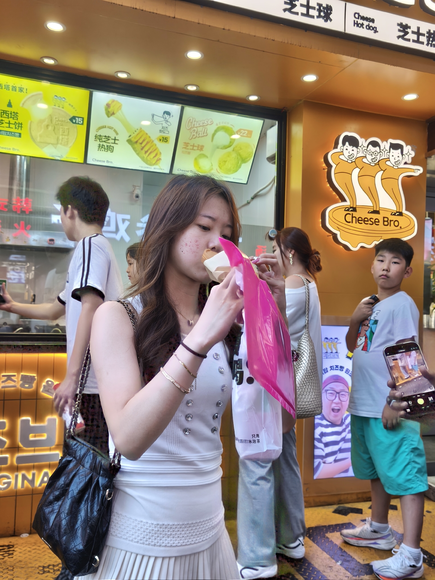A young East Asian woman is captured mid-bite in a street food setting in Heping District, China. She is wearing a white sleeveless top adorned with rhinestones and a pleated white skirt. A black quilted handbag with a chain strap rests on her left hip. She holds a food item wrapped in a beige paper cone, partially obscured by a bright pink plastic bag. Her right hand is visible, holding the pink bag. Her hair is long and brown, falling over her shoulders.
Behind her, a young East Asian boy in a white t-shirt and teal shorts stands with his arms crossed, holding a smartphone in his right hand, which displays an image. To the boy's left, another young East Asian woman, wearing a white top and a large metallic tote bag, walks away from the camera. Further back, a young East Asian man in a white t-shirt with black stripes is seen from the back.
The background features a storefront with bright yellow signage and illuminated menu boards displaying images of food, including cheese-filled items like hot dogs and balls. A prominent logo features three cartoon characters with the text "Cheese Bro." The ground is a patterned surface, possibly a sidewalk, with yellow and dark striped lines. The overall scene suggests a lively street food vendor area.
