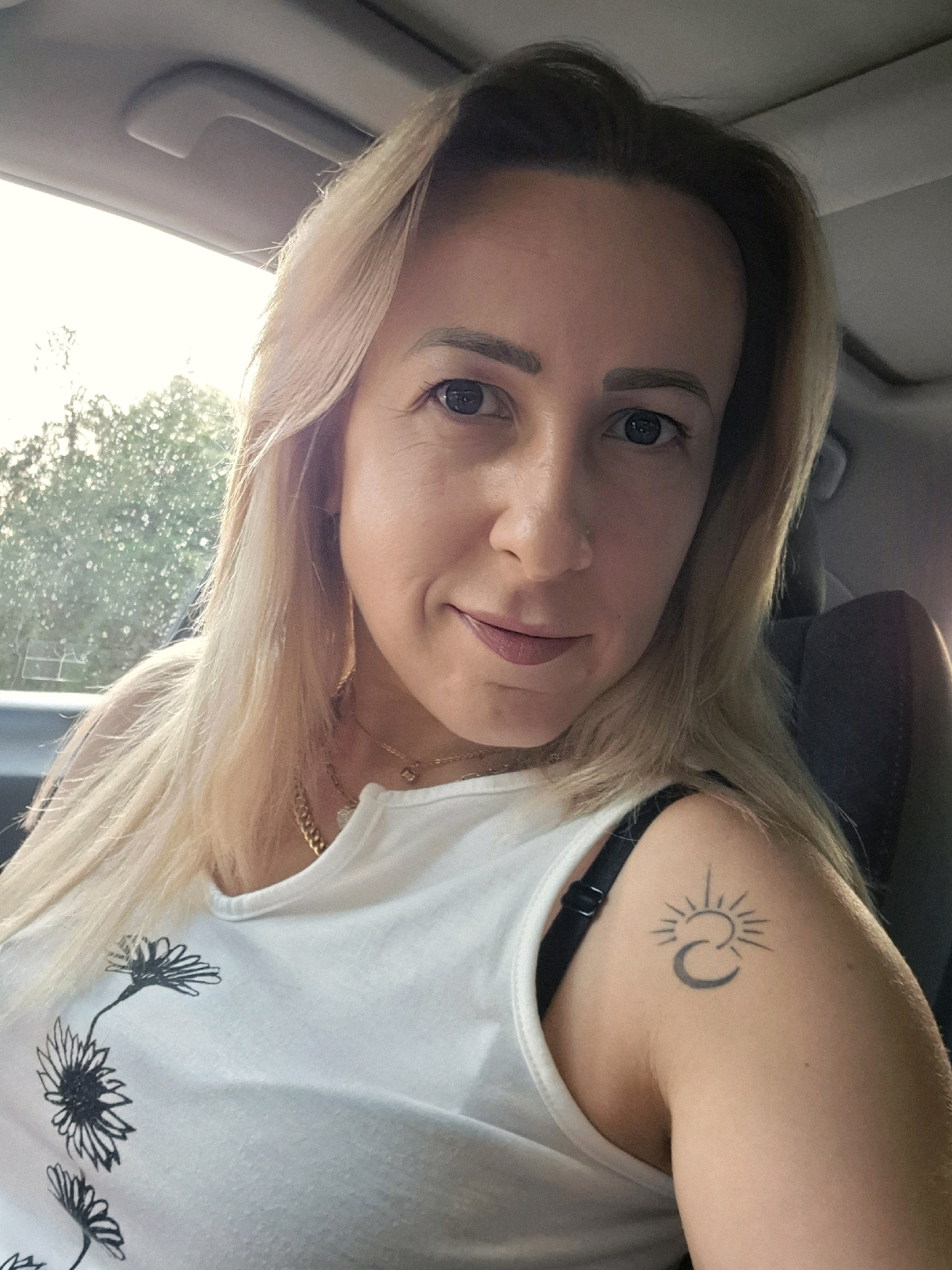 A fair-skinned blonde woman is captured in a selfie from the chest up, inside a vehicle. She has blue eyes, light brown eyebrows, and is wearing a white tank top with a floral design. A black bra is visible beneath the tank top, and she has a tattoo on her left shoulder featuring a sun and moon design. She is also wearing a gold chain necklace with a small pendant. The background shows the interior of a car, with sunlight streaming in from the left, illuminating trees outside the window. The overall lighting is bright, with some areas of shadow.