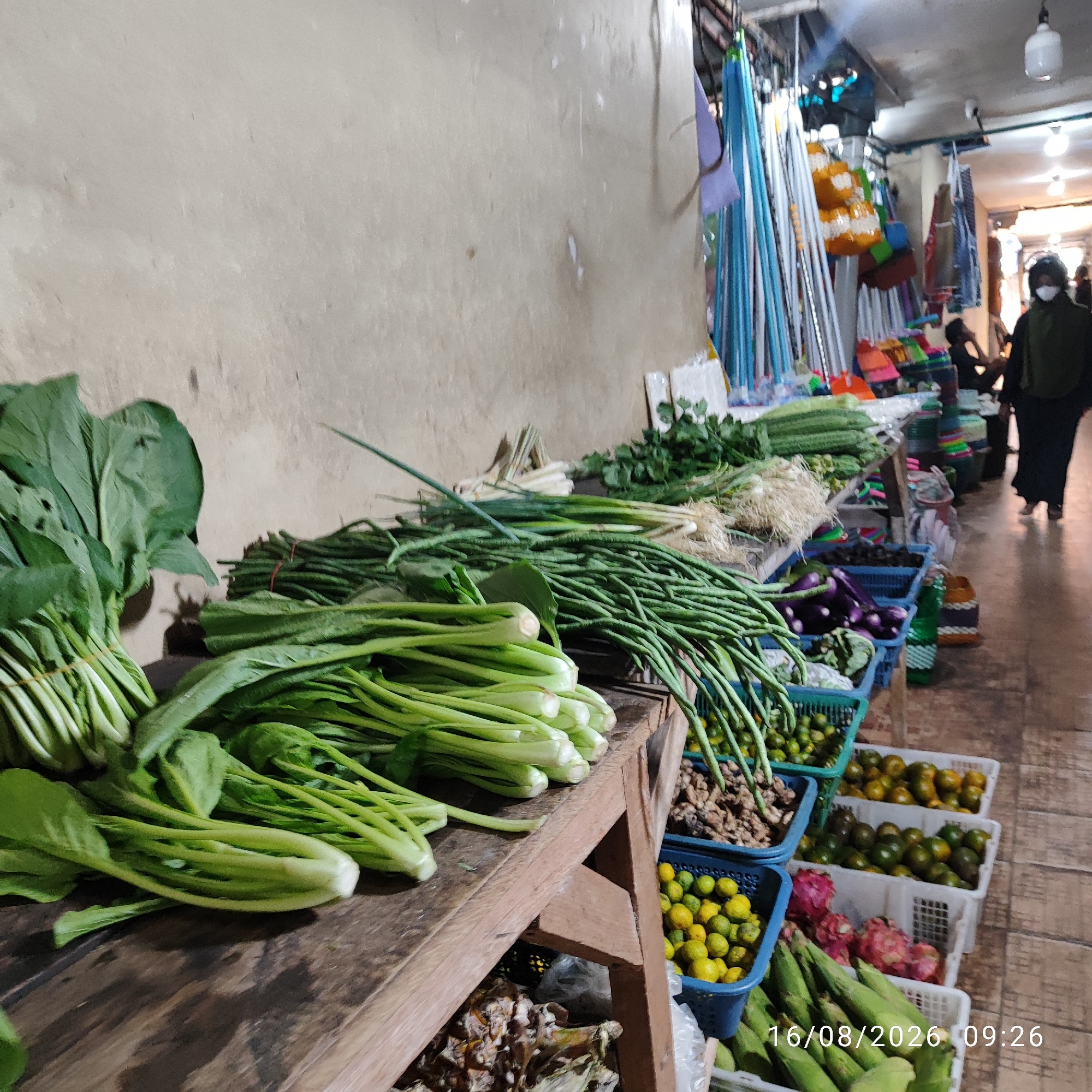 The image shows a street market scene in Pembaharuan, Indonesia. In the foreground, a wooden stall displays an assortment of fresh vegetables, including leafy greens like mustard greens and what appear to be bok choy or similar Asian greens, as well as long green beans. Behind the vegetables, other produce is visible in plastic baskets, such as eggplants, small citrus fruits (likely limes or calamansi), and oranges. In the background, a person wearing a dark hijab and a mask walks away from the camera down a narrow pathway lined with stalls. These stalls exhibit various goods, including what seem to be cleaning supplies like brooms and mops, and colorful umbrellas. The environment appears to be an open-air market with a concrete floor and a covered ceiling with lighting fixtures. A timestamp in the bottom right corner reads "16/08/2026 09:26".