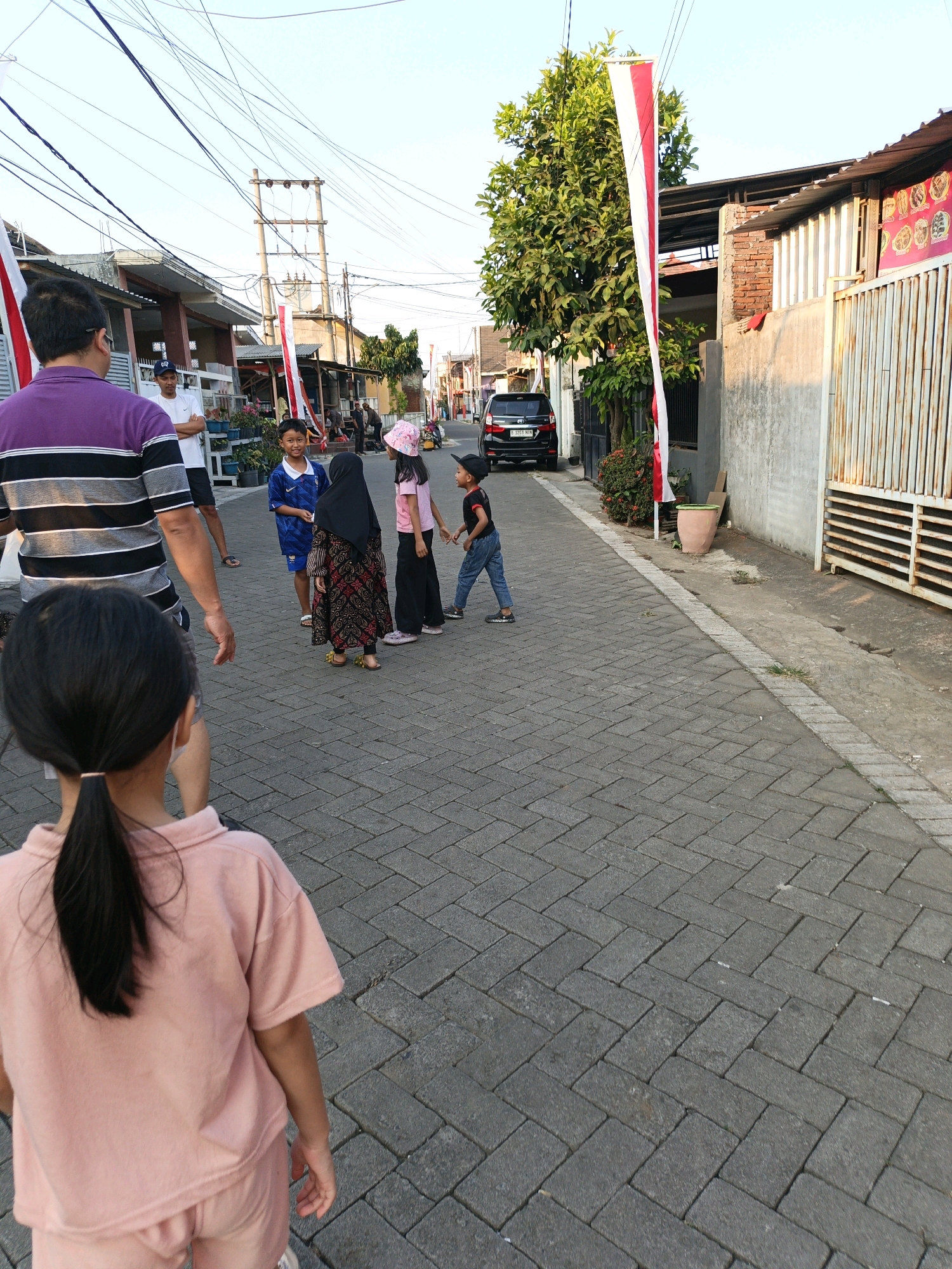 In Karangploso, Indonesia, a street scene unfolds with several individuals in an outdoor environment. The street is paved with interlocking gray bricks arranged in a herringbone pattern. The sky above is clear and bright. On the left side of the street, a man wearing a purple and black striped shirt stands with his back to the camera. Further down the street, a group of children are visible. One boy in a blue soccer jersey looks towards the camera. A girl wearing a black hijab and a patterned dress is also present, along with another girl in a pink hat and shirt. A boy in a black shirt and blue jeans is walking away from the group. In the background, a black car is parked on the side of the road. Buildings line the street, some with red brick accents and metal fencing. Red and white Indonesian flags are displayed vertically on poles, adding a festive touch. A tall tree with lush green leaves stands prominently on the right side of the street. The overall impression is of a casual, communal gathering in a residential urban setting.