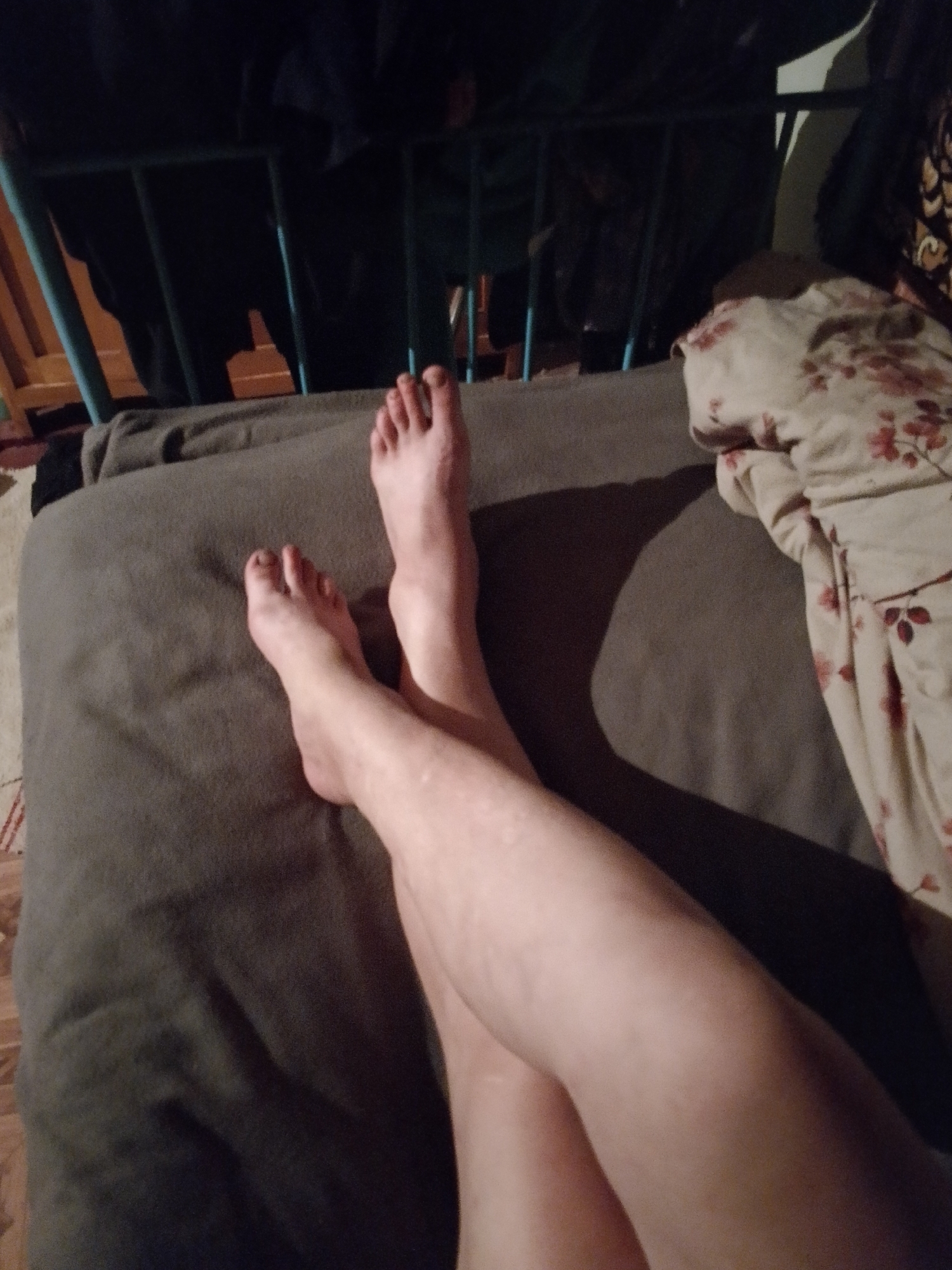 The image displays a downward-facing perspective of a person's bare legs and feet, positioned on a grayish-brown upholstered surface. The feet are visible in the upper center, with the legs extending diagonally towards the lower right, appearing to be crossed. The skin tone is fair. Toenails on both feet are painted with a dark pigment. A distinct shadow is cast by the left leg onto the upholstered surface. To the right, a crumpled light-colored blanket or textile featuring a red and brown floral pattern is visible. The upper left background reveals a dark, indistinct area with a partial view of a blue-green metal or wooden frame structure. The overall lighting is dim, suggesting an indoor environment. The location is Unknown, Unknown.