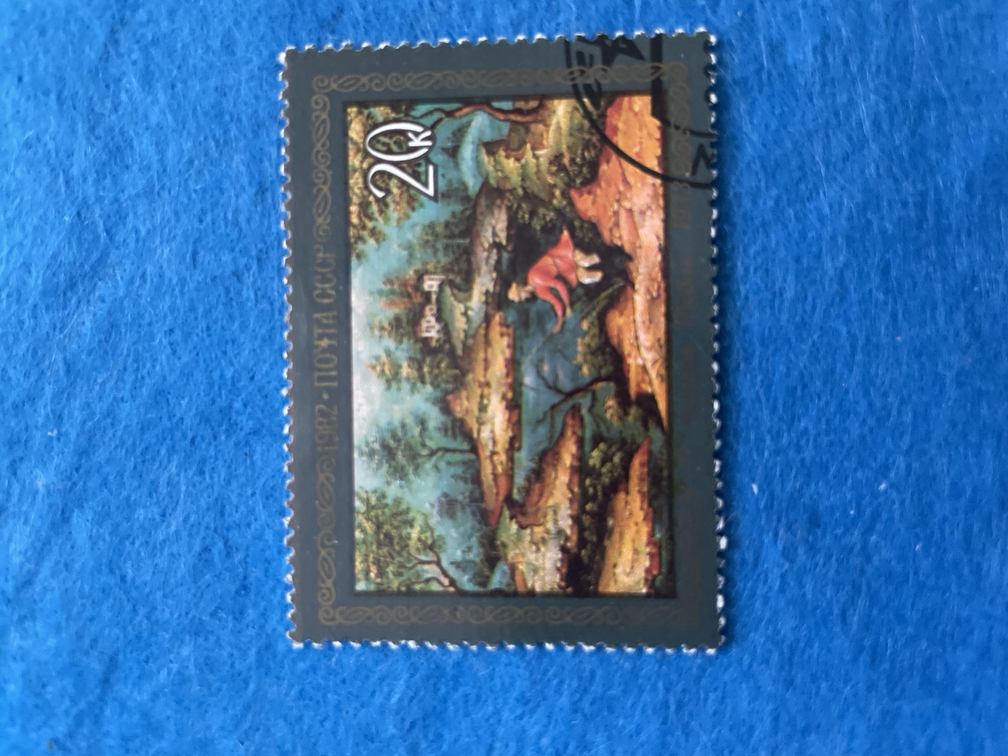 This is a close-up, high-angle shot of a postage stamp against a textured blue background. The stamp features a painting of a pastoral landscape. In the foreground, a person in a red jacket and white pants is depicted fishing by a body of water. The background includes trees, a house, and a body of water. The stamp has a serrated edge and text that reads "1982 ПОЧТА СССР" and a numeral "20" with a small "K" below it. The blue background has a fuzzy, carpet-like texture. There is no indication of the city of Perm, Russia, in the image.