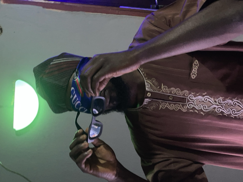 A person wearing a dark-colored cap and a blue Gucci face mask holds up a pair of sunglasses. The individual is dressed in a brown, intricately embroidered shirt. A green, semi-circular light fixture illuminates the scene. The image is oriented in portrait format, but the subject is tilted to the right.