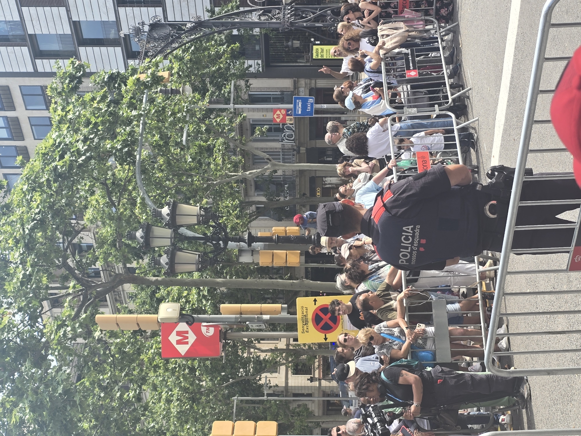 An outdoor, daytime scene on a city street in Barcelona, Spain, features a crowd gathered behind a metal barricade. A police officer in uniform, with "POLICIA mossos d'esquadra" visible on their back, stands facing the crowd. In the background, people are observing something, some holding phones. Various street signs are present, including one with the "M" logo for the Barcelona Metro and a sign indicating "Sanyalitzacio Excepcional" (Exceptional Signage). A bus stop sign is also visible. Tall trees line the street, and buildings with architectural details are in the background. The overall atmosphere suggests a public event or procession.